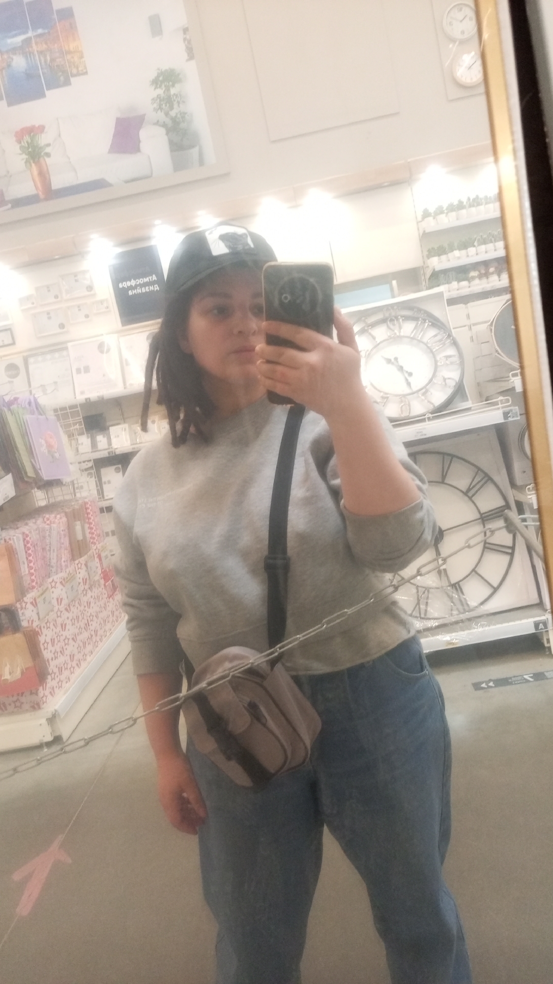 The image depicts a woman taking a selfie in a store setting. She is wearing a gray sweatshirt, blue jeans, and a baseball cap. A small gray bag hangs across her body. The background reveals store shelves stocked with various items, including clocks and home decor products. Based on additional information, the location is Юдино, Russia.