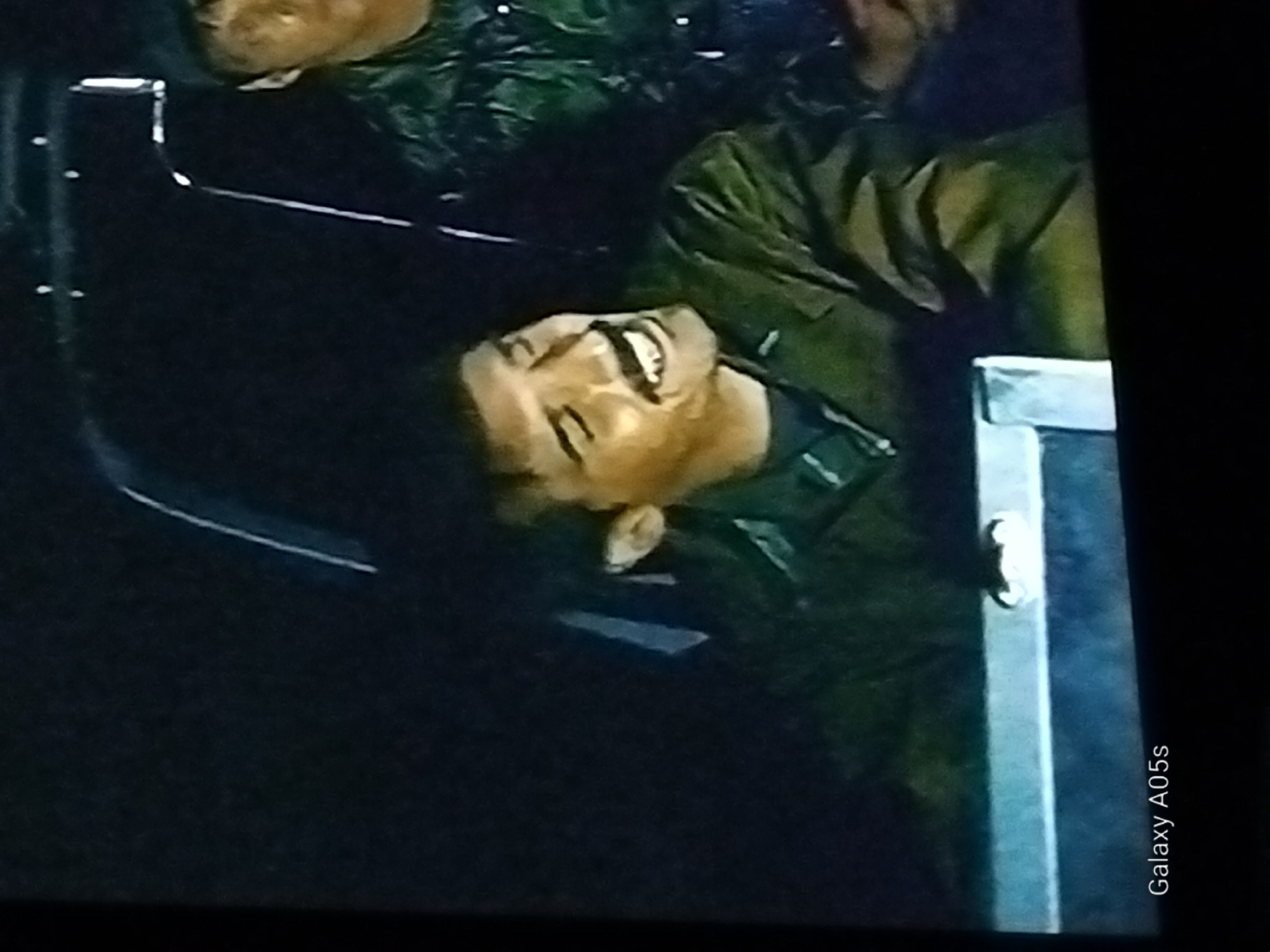 The image depicts two individuals inside a vehicle at night. The person in the foreground is a man with dark hair, a prominent mustache, and is smiling broadly, showing his teeth. He is wearing a dark green jacket over a dark shirt. The man in the background, to the right, is wearing a dark cap and a camouflage-patterned garment. The interior of the vehicle is dimly lit, with some light reflecting off a metallic surface in the foreground. The "Galaxy A05s" watermark is visible at the bottom left corner. There is no discernible indication of the location being the city of Druzhba, Russia.