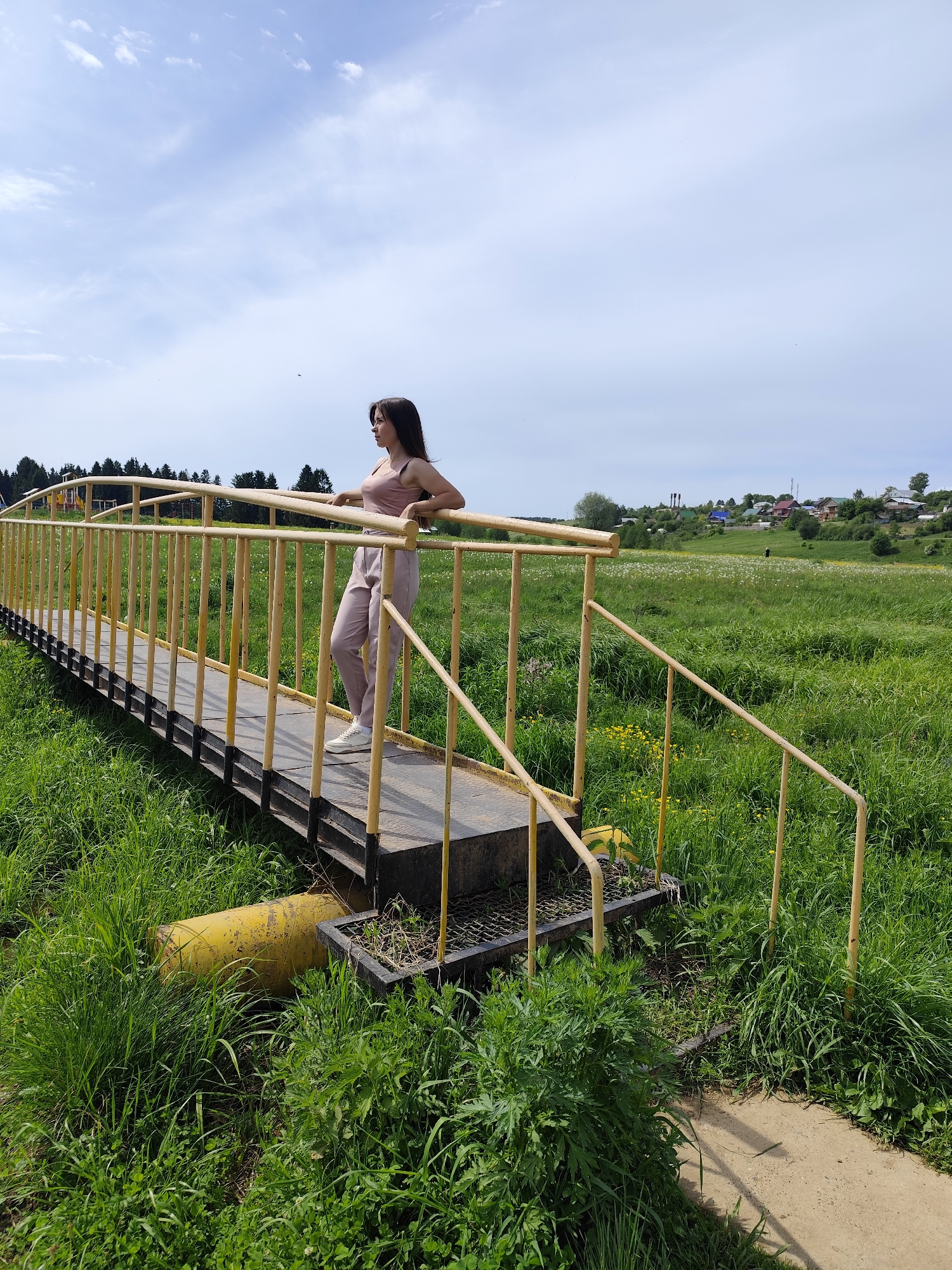 A medium, full outdoor shot features a young woman standing on a small yellow bridge in a field of tall green grass, likely in the vicinity of Фоки, Russia.
The woman, facing left, rests her hands on the railing. She has long dark hair and is wearing a mauve tank top and matching pants with white sneakers. The small bridge is painted yellow with a black platform and grated metal at the ends. The surrounding field is full of lush green grass, punctuated by patches of white and yellow wildflowers. In the background, a row of trees and some houses can be seen. The sky is light blue with scattered white clouds. The lighting suggests it is daytime. The overall impression is one of a peaceful, rural scene.