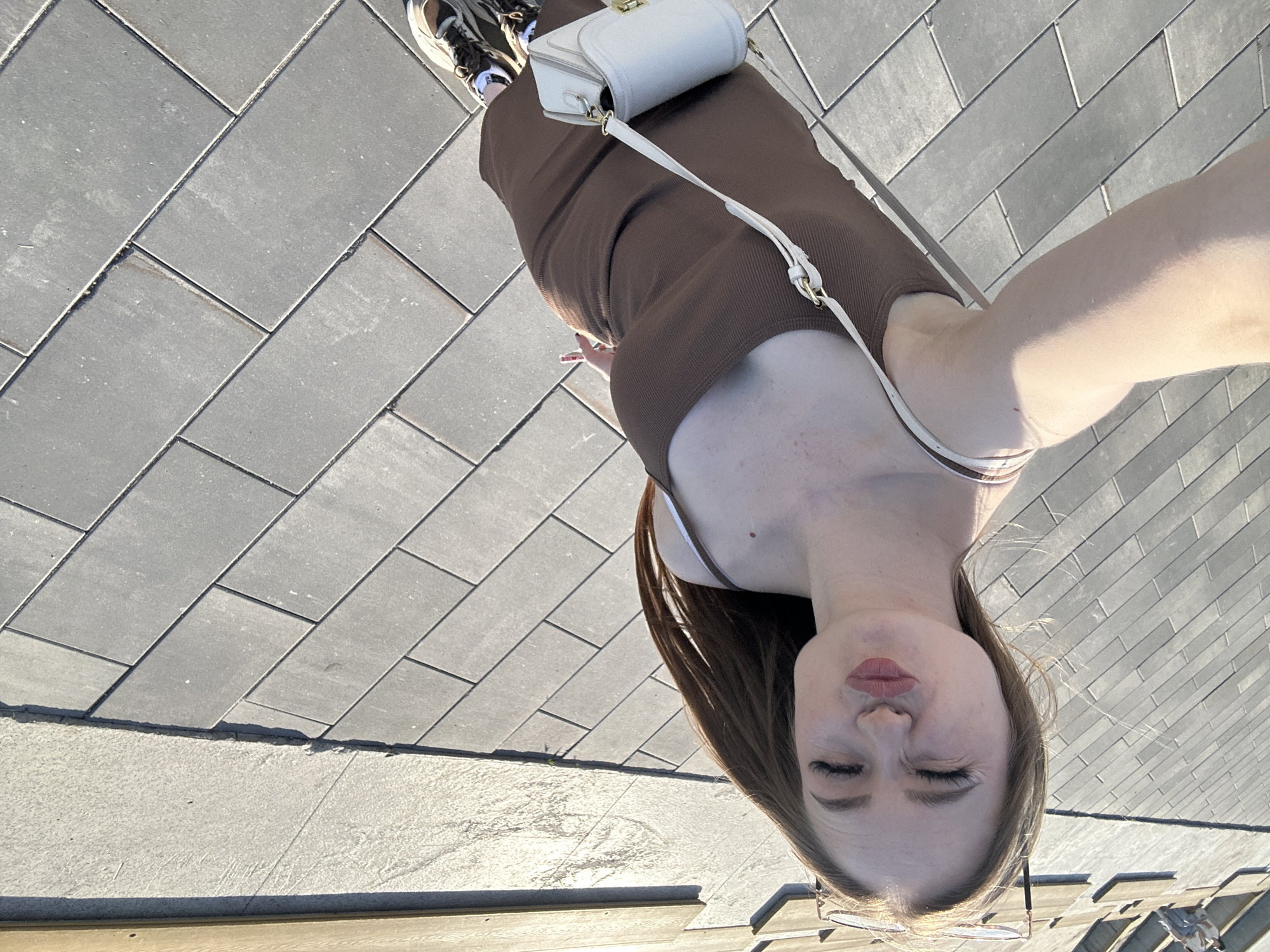 The original image depicts a young woman in an unusual, rotated perspective, appearing upside down with her head at the bottom of the frame and her feet towards the top, looking down at the camera. She has long, dark brown hair, and her eyes are mostly closed in a squint, with her lips pursed in a slight pout. Clear-framed glasses rest on her nose.
She is wearing a dark brown, ribbed, sleeveless top or dress, with a white bra strap visible on her left shoulder. A white structured handbag with a gold clasp and a thin strap is slung across her body. Her feet, visible in the upper right, are clad in sneakers with dark and lighter, possibly metallic, elements.
The woman is standing on a grey paved surface, composed of rectangular interlocking pavers laid in a diagonal pattern. To her right (the left side of the frame), there is a strip of lighter, textured concrete or similar material, bordering the pavers. A wooden or composite board runs horizontally along the bottom edge of the frame.
The scene is brightly illuminated by direct sunlight, indicated by strong highlights and distinct shadows, suggesting a midday or early afternoon setting. There are visible freckles and a small mole on her chest/shoulder area. No visible text is present in the image.