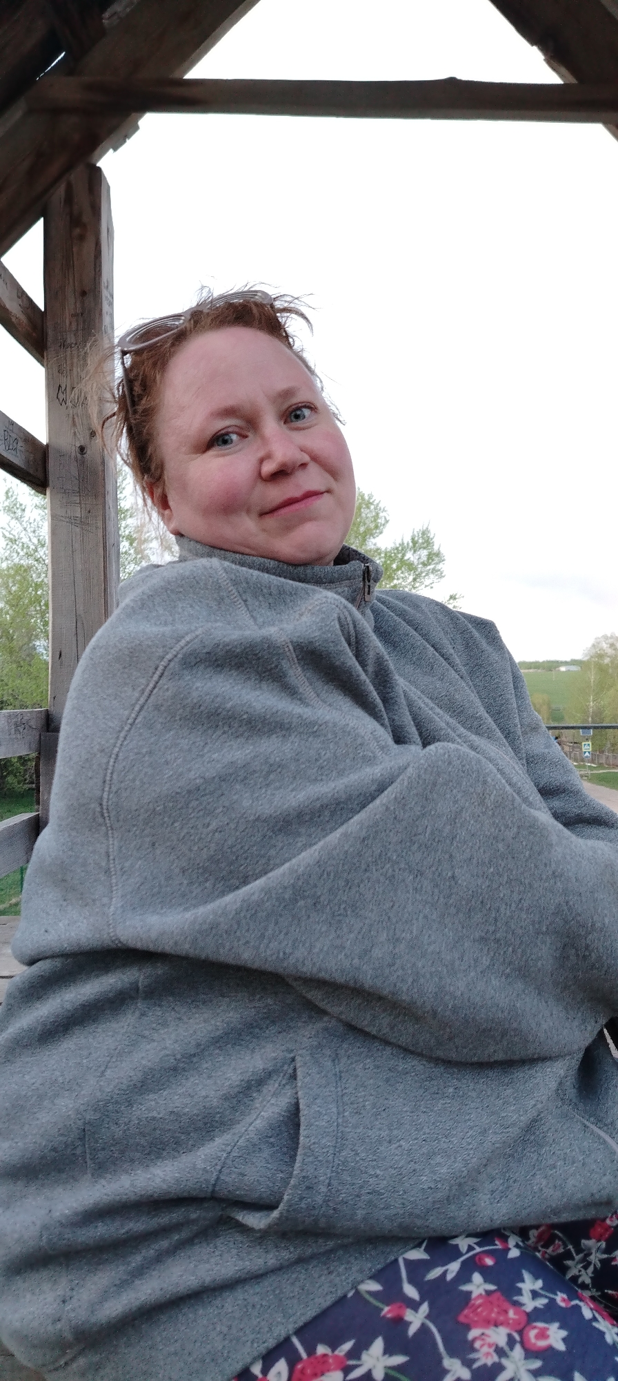A medium shot depicts a middle-aged woman with fair skin and reddish-blonde hair sitting outdoors in Зипуново, Russia. She wears a gray fleece jacket and floral print pants. The woman is seated within a wooden structure, possibly a gazebo, with supporting beams visible behind her left shoulder. Overhead, the wooden roof of the structure is visible. Sunglasses are perched on top of her head. The background features green trees and an open landscape.