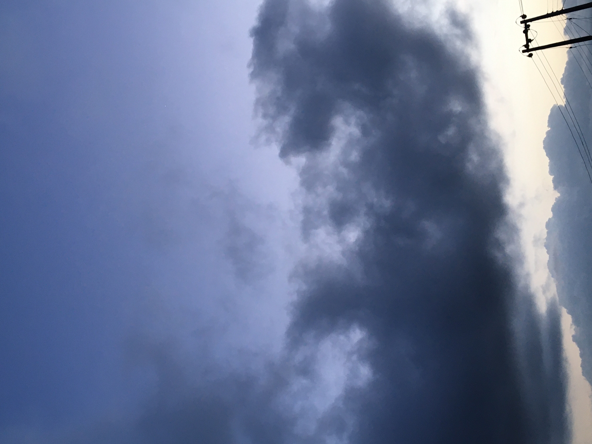 The image depicts a dusk sky over Agege, Nigeria. Dark, heavy clouds dominate the upper half of the frame, obscuring much of the sky and creating a moody atmosphere. Towards the bottom of the image, lighter, wispy clouds are visible, interspersed with the silhouettes of utility poles and their associated wires. The sky transitions from a deep, moody blue at the top to a pale, almost white hue near the horizon, suggesting the last vestiges of daylight. The scene is devoid of people and any discernible activity, presenting a static, environmental portrait of the sky at twilight in this Nigerian locale.