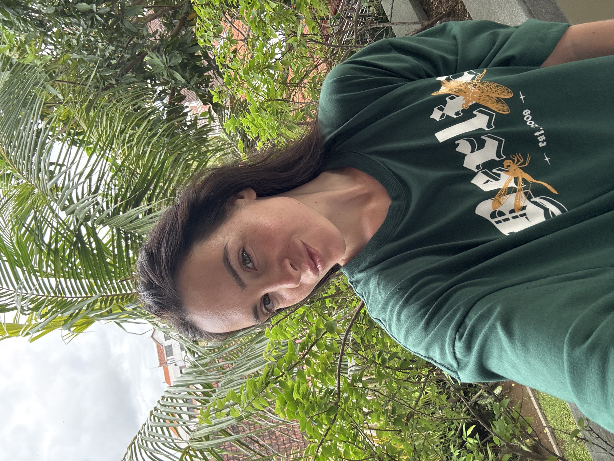 A young Caucasian woman with long, dark brown hair is taking a selfie outdoors. She is wearing a dark green long-sleeved t-shirt with white text and two orange dragonfly graphics. Her head is tilted to her right, and she is looking towards the camera with a slight smile. The background features lush green foliage, palm fronds, and a building with a red tile roof, indicative of North Kuta, Indonesia. The sky is overcast.