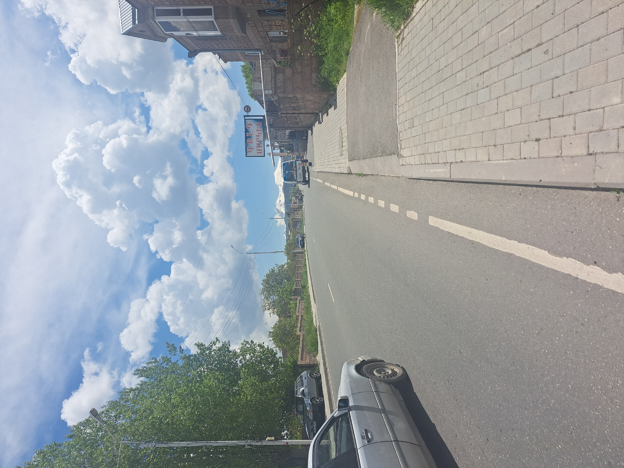 This image captures a street scene in Artik, Armenia, on a clear, sunny day with a bright blue sky partially filled with white cumulus and cirrus clouds. The perspective is from the driver's side of a light-colored vehicle, looking down a two-lane asphalt road.
In the immediate foreground on the left, a silver/grey Mercedes-Benz sedan is partially visible, showing its front left wheel and part of its body. The asphalt road surface is visible, marked by a dashed white line separating traffic lanes and a solid white line defining the shoulder or parking area on the right.
Along the left side of the road, a dense line of lush green trees partially obscures the background. A grey utility pole with overhead wires is prominent on the left. Further down the left, a grey hatchback vehicle, possibly a Lada Samara, is parked facing away from the viewer, with its license plate "64 ZC 010" partially readable. Behind it, a low stone wall and more trees are visible.
On the right side of the road, a paved sidewalk with light grey rectangular pavers runs parallel to the street, separated by a concrete curb. A stone wall, constructed from light brown or reddish-brown blocks, extends along the right side, bordering properties. Beyond this wall, a building with a distinctive bay window and a metal roof is visible. Further down the road, a sign with Armenian text "ԳՈՒՄ ԱՐՏԱԿ" (GUM ARTAK) is visible above the stone wall, likely indicating a business.
A white van, possibly a Ford Transit, with "Mole Tours" visible on its side, is driving away in the distance on the right lane. Behind the van, snow-capped mountains are visible in the far background under the clear sky. Several other utility poles and power lines crisscross the scene, particularly above the road and buildings on the right. Sparse green vegetation, including grass and weeds, grows along the edges of the sidewalk and walls. No people are discernible in the scene.