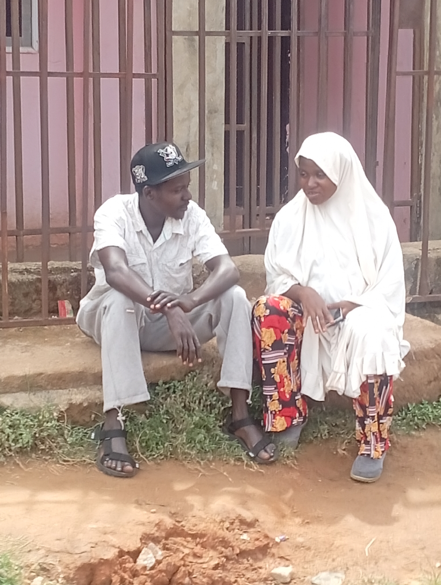Two people are sitting on a concrete step outside a building with pink walls and metal bars in Sabon Tasha, Nigeria. A man wearing a black baseball cap, a light-colored short-sleeved shirt, gray pants, and black sandals is seated on the left. He is looking towards a woman seated on the right. The woman is wearing a white hijab and a white dress over colorful patterned pants. She is holding a mobile phone. The ground is brown dirt with some patches of grass.