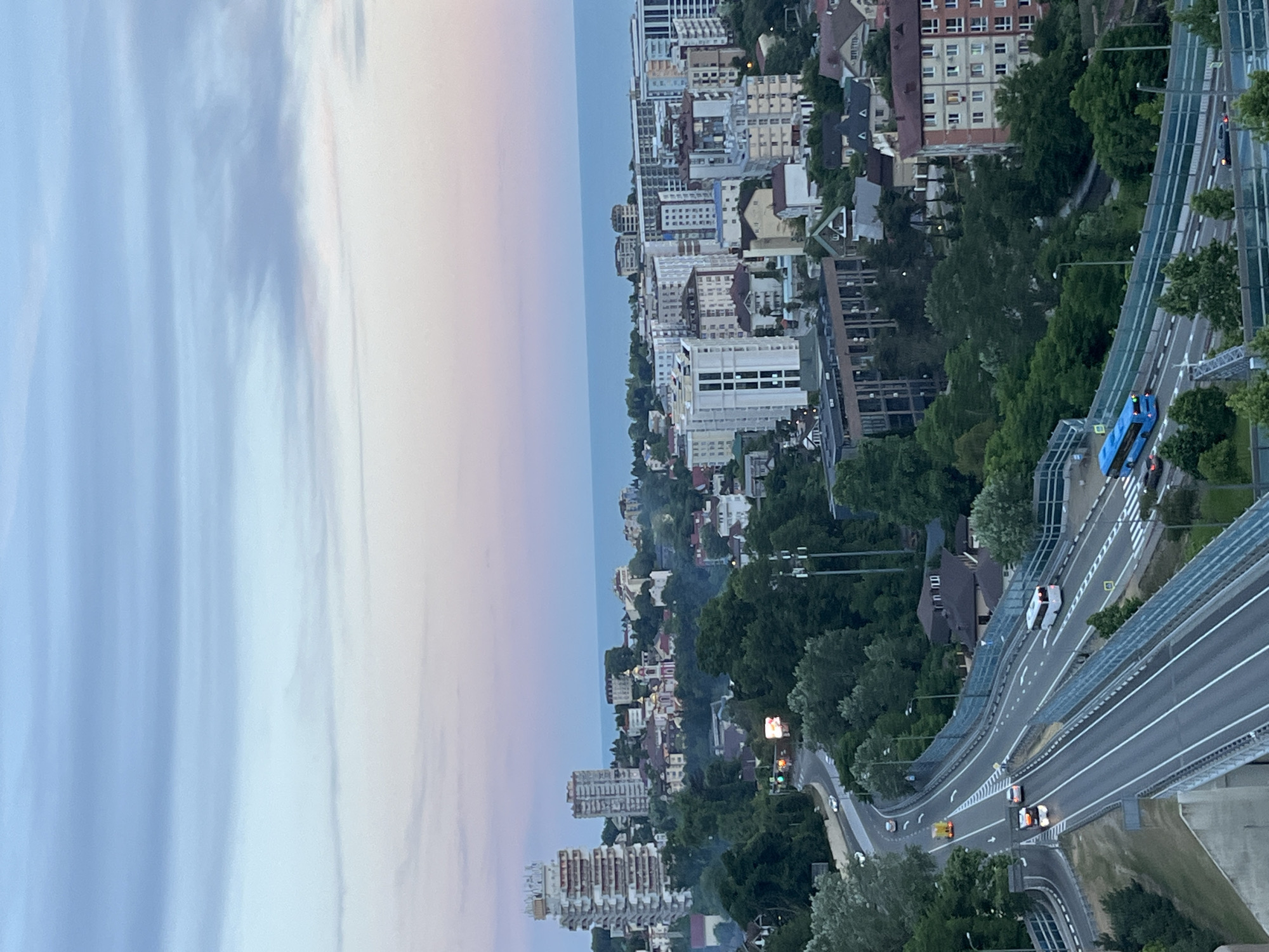 This is a high-angle, medium shot of a cityscape and highway in Sochi, Russia, near sunset. The sky is a gradient of pale blue and pink. In the background, modern buildings and a body of water are visible. A highway curves through the foreground, with several vehicles, including a white bus and a blue bus, traveling on it. The highway is lined with trees and fences.