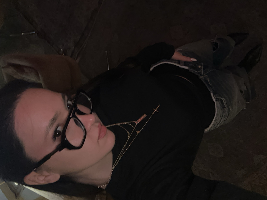 The image presents a female individual in a high-angle, full-body self-portrait. She possesses dark hair and wears black-framed eyeglasses. Her attire includes a long-sleeved black top, light-wash distressed denim jeans, and dark pointed-toe footwear. Multiple layered necklaces adorn her neck, one featuring a rectangular pendant and another with a cross pendant. Her right hand, exhibiting light-colored nail polish, is partially inserted into a jeans pocket. The background is dimly lit and largely out of focus. Observable elements include a light-colored, possibly upholstered object in the upper right quadrant and a dark, textured floor or rug. Faint vertical metallic rods are visible on the right edge. The specific location within Yerevan, Armenia, is not ascertainable from the visual information provided.