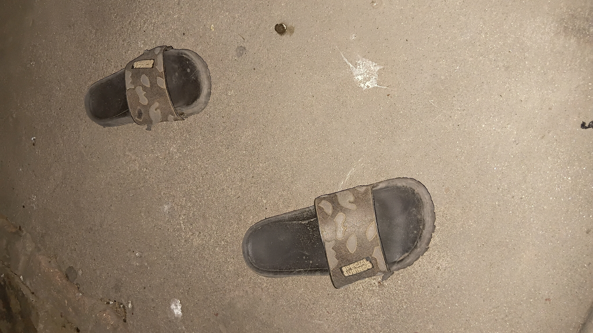 Two camouflage-patterned slides with black soles are on a concrete surface. The slide on the left has "SPECIAL SUBING" text visible on the strap. The surface appears gritty and shows some debris and white splotches, possibly bird droppings. The environment suggests an outdoor urban setting, potentially in a less maintained area of Prabumulih, Indonesia.