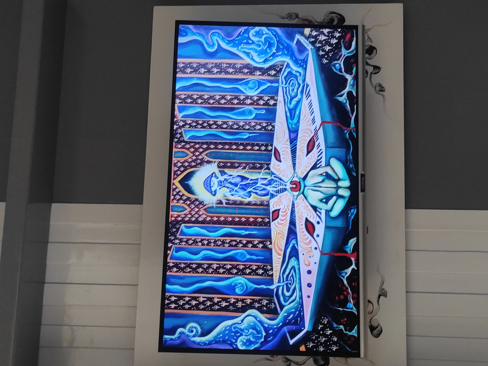 The image shows a television screen displaying a colorful, surreal artwork. The artwork features a central figure with a red eye, seated in a lotus-like pose, with a glowing mushroom-shaped entity above its head. The background consists of ornate columns and swirling blue patterns. The television is mounted on a wall with vertical white panels on the left and a dark gray panel on the right. The bottom of the screen shows a metallic strip, possibly the TV's bezel or a soundbar. Below the television, decorative swirling motifs are visible on the wall. The overall scene is an indoor setting, likely a room, but the specific location within Sumedang, Indonesia, cannot be determined from the provided image.