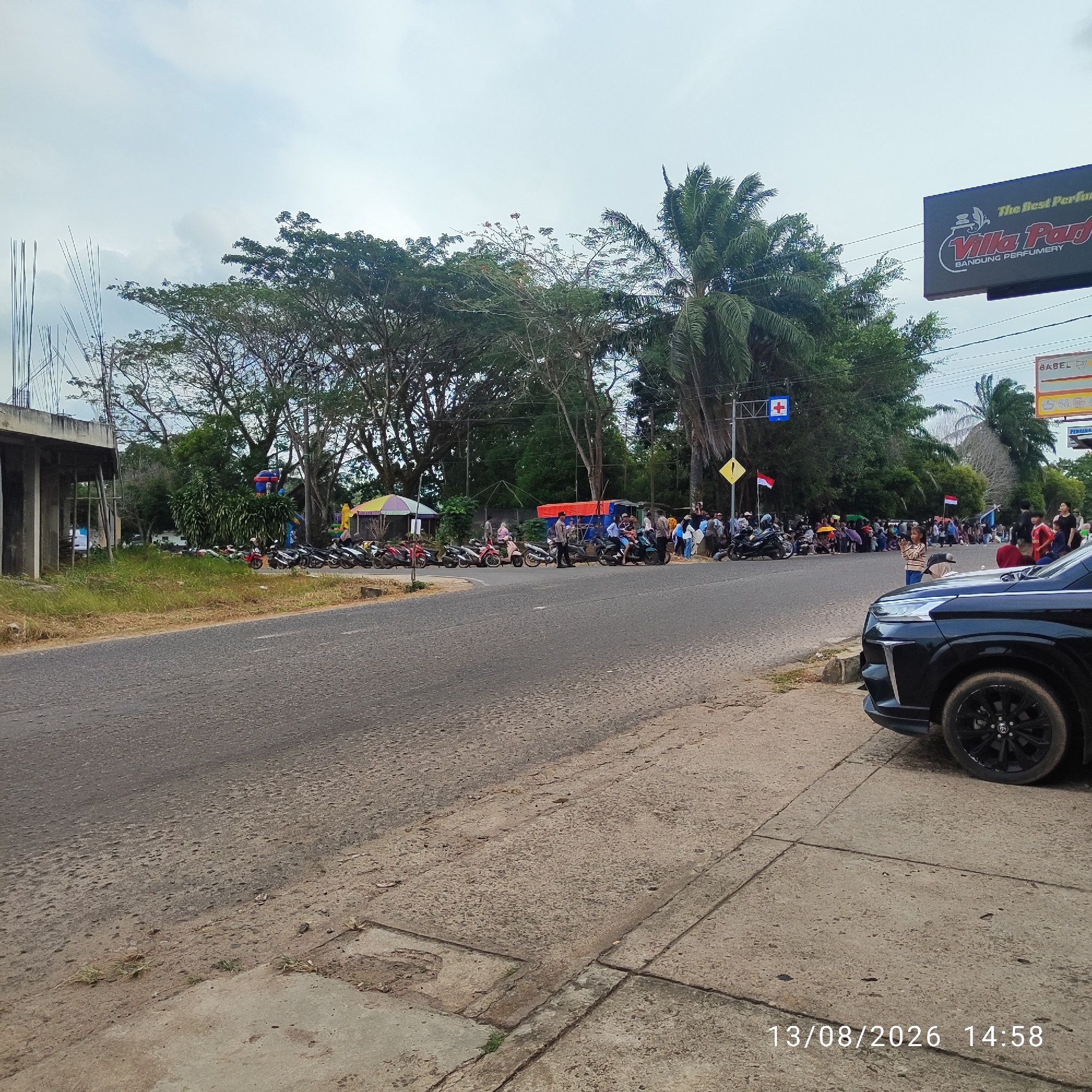 The image depicts a street scene in Buding, Indonesia, on August 13, 2026, at 2:58 PM. A black car is parked on the right side of the frame, partially obscuring the sidewalk. The street itself is paved with asphalt and appears to be in moderate condition with some cracks and unevenness.
On the left side of the street, there is an unfinished concrete structure with rebar protruding upwards, suggesting ongoing construction. Adjacent to this structure, grass and vegetation grow along the roadside.
Across the street, a line of parked motorcycles is visible. Beyond the motorcycles, a crowd of people is gathered. Some individuals are standing, while others are seated, and many appear to be holding umbrellas for shade. Several colorful stalls or awnings are set up, with one featuring a red tarp and another with a yellow and pink striped umbrella.
In the background, there are numerous trees, including palm trees and other deciduous varieties, creating a lush, green environment. A billboard with the text "The Best Perfumery Villa Park BANDUNG PERFUMERY" is prominently displayed on the right side of the image, above the trees. Traffic signs, including one with a red cross symbol and another with a yellow warning sign, are also visible along the street. Two Indonesian flags are planted near the gathered crowd. The sky is overcast with scattered clouds.