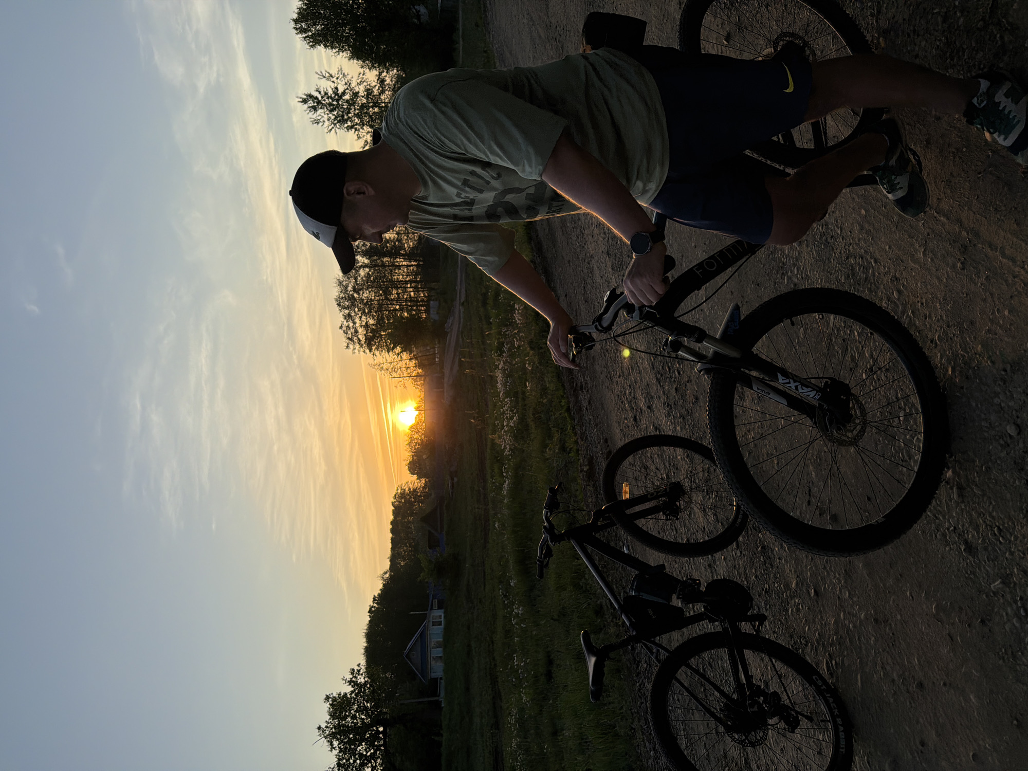 A man wearing a baseball cap, green t-shirt, and blue shorts is riding a bicycle on a dirt road in Noviy, Russia, at sunset. The sun is low on the horizon, visible through the trees. There is another bicycle parked next to him. The setting is rural with some trees and a building visible in the background.