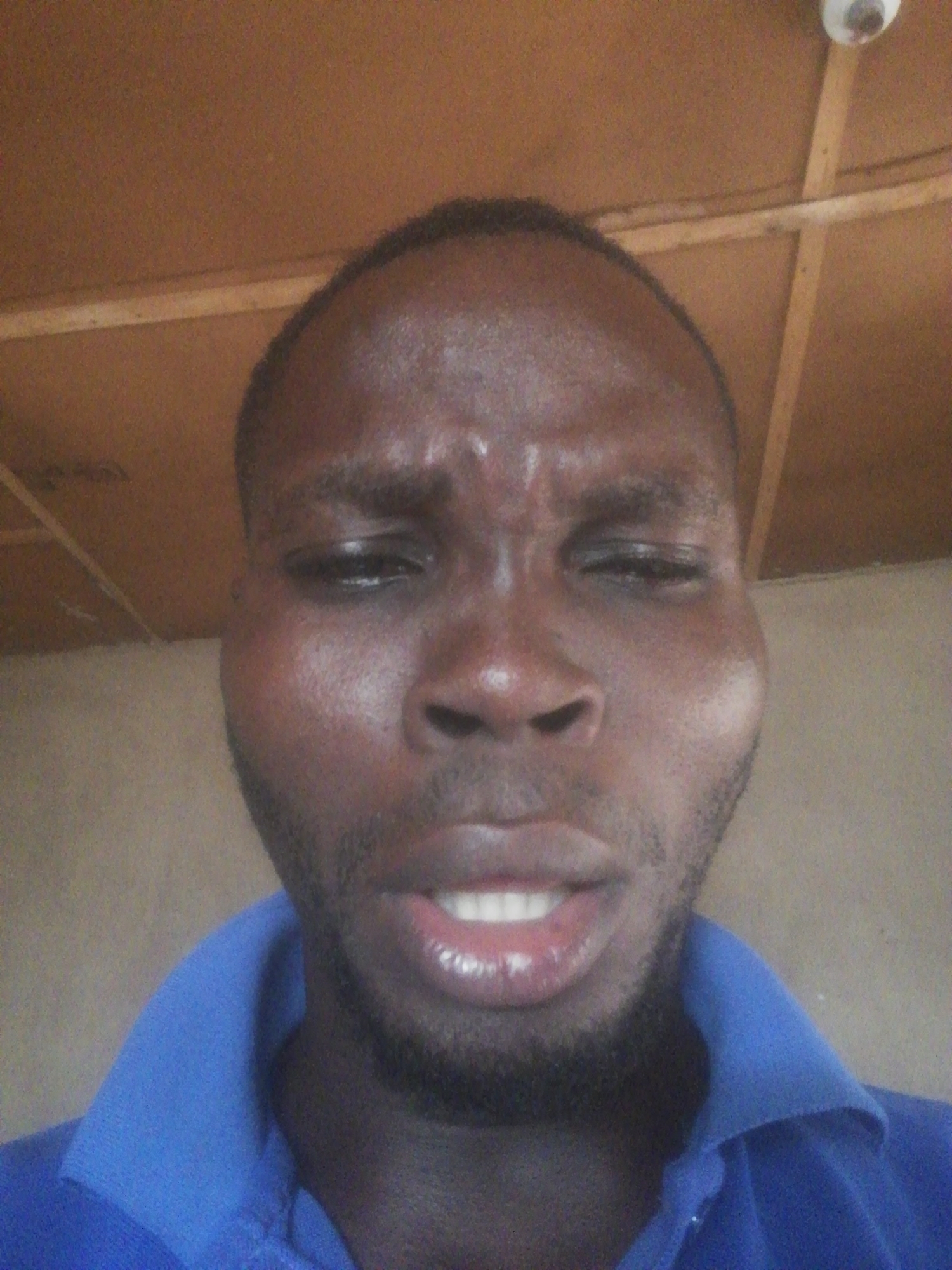 A close-up selfie of a Black man with a short haircut and beard, wearing a blue collared shirt. He has a furrowed brow and a slight grimace. The background consists of a wooden ceiling and a wall. The location is Unknown, Unknown.
