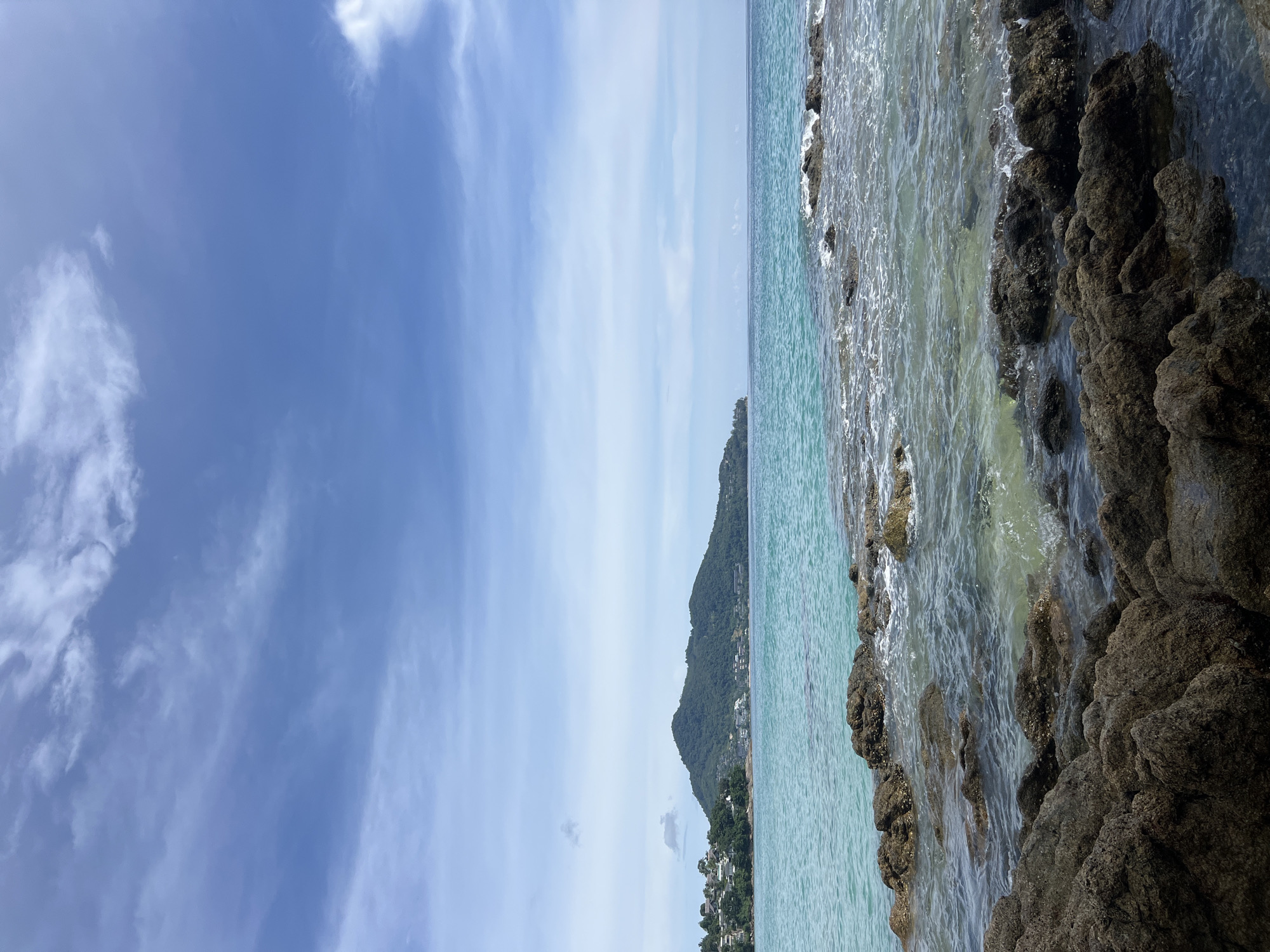 This is a vertical, eye-level, outdoor, medium-shot photograph taken on a sunny day at a beach in Choeng Thale, Thailand. The image depicts the turquoise waters of the ocean meeting a rocky shoreline. The sky is a clear blue with scattered white clouds, suggesting fair weather.
The scene is dominated by the natural elements of the sea and sky. The ocean's surface is calm with gentle ripples, and the water is clear enough to reveal the submerged rocks beneath. The foreground is occupied by rugged, dark brown rocks that are wet from the waves. In the background, a lush green, tree-covered mountain slopes down towards the coast. Dotted along the base of the mountain, near the water's edge, are buildings, suggesting a populated coastal area or town. The overall mood is serene and tranquil, evoking a sense of natural beauty and peace. There is no visible text in the image.
