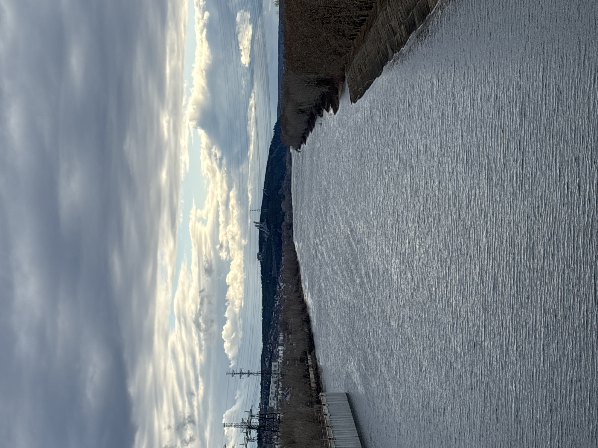 The image displays a wide, calm body of water, likely a river, extending horizontally across the frame. Its surface is a uniform, muted grey, covered with numerous small ripples, suggesting either a slight current or reflections of an overcast sky.
The sky occupies the left half of the image, filled with expansive, layered grey clouds. Brighter, whiter clouds and hints of a paler blue sky are visible in patches, particularly towards the right side of the sky, indicating a partially overcast day.
Along the bottom edge of the water, a riverbank is visible, densely covered with bare, dark-branched trees and shrubs, characteristic of late autumn, winter, or early spring. Several large, dark metal power line towers, with multiple lines stretching across the landscape, are prominent features on this bank. A long, grey industrial-looking dock or platform extends into the water from this near bank. In the mid-distance along this bank, obscured by trees, some buildings of a town or settlement are discernible.
The opposite bank, positioned along the top edge of the water, also features a strip of dense, leafless trees and vegetation, behind which the land rises into a forested hill or ridge.
The scene depicts a static natural landscape, interspersed with human infrastructure. No people or specific activities are visible, and there is no discernible text. The overall impression is one of a vast, tranquil waterscape within a developed region, consistent with the provided context of Chaikovsky, Russia, and potentially the Kama River.