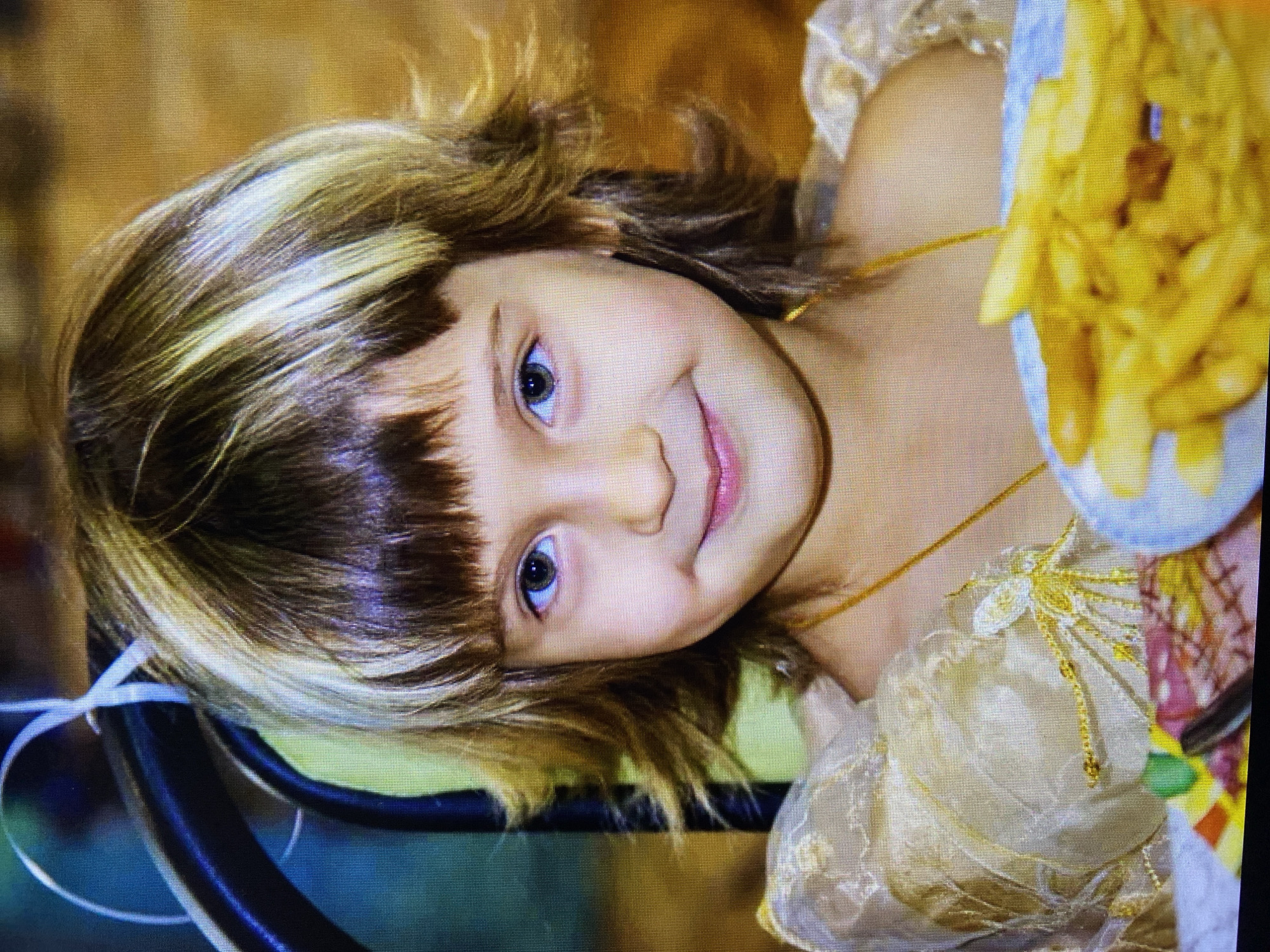 A young girl is captured in a close-up, waist-up shot, seated at a table. Her hair is styled in a layered cut with bangs, featuring streaks of blonde and brown. She has striking blue-green eyes and a gentle smile. She is wearing a gold-colored dress with ornate embroidery on the sleeves and a thin gold necklace. In the foreground, a plate filled with french fries is visible, suggesting a meal is being enjoyed. The background is softly blurred, with hints of warm colors and some greenery, possibly indicating an indoor or outdoor setting. The image appears to be a candid photograph, focusing on the girl's cheerful expression.
