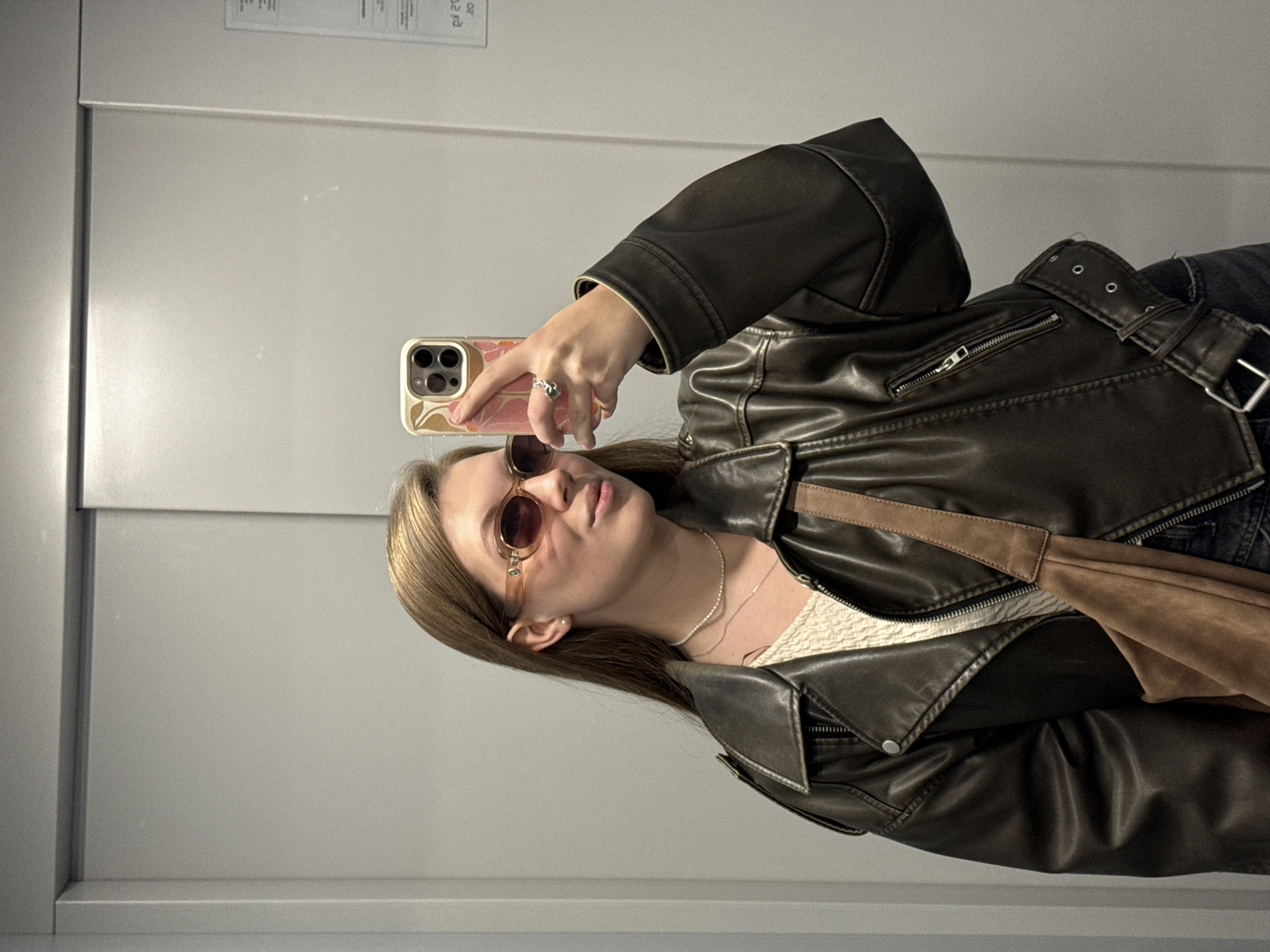 A young woman with long brown hair, wearing brown-tinted sunglasses, a brown leather jacket, and a light-colored top, takes a selfie. She is holding a mobile phone with a patterned case. The background is a plain, light gray wall. The photo appears to be taken indoors, possibly in an elevator. No geographical location is discernible from the image.