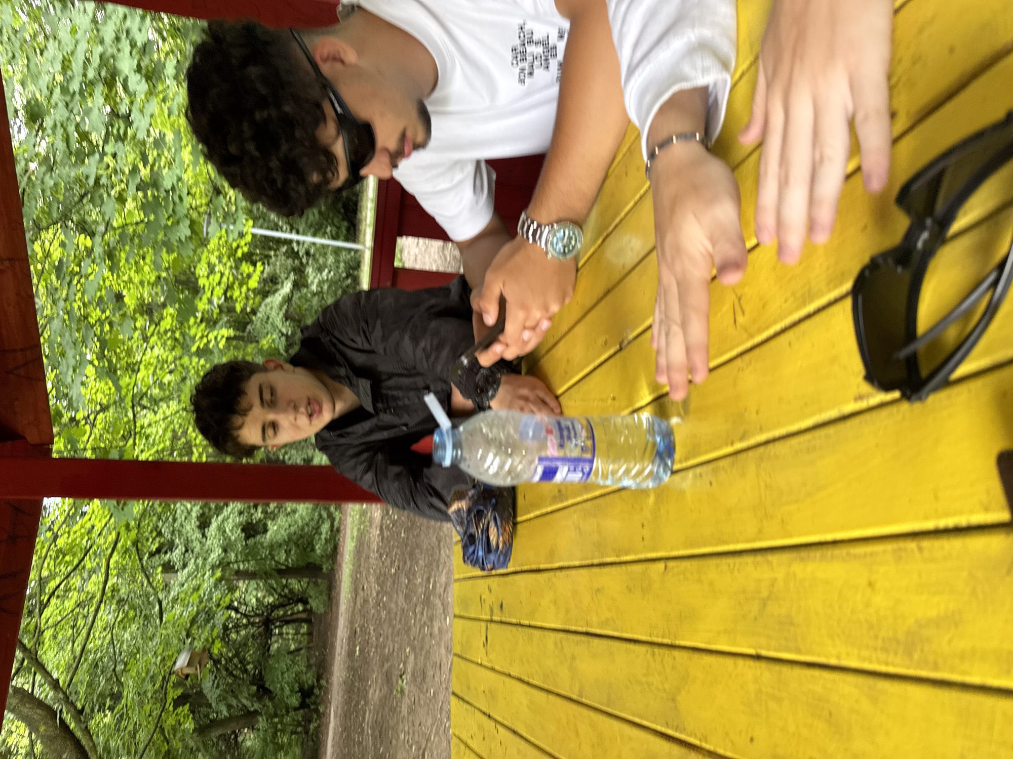 The image captures an outdoor scene featuring three individuals at a bright yellow wooden table, likely within a gazebo or open-air shelter with red structural beams. The background is lush with vibrant green foliage and trees, and a dirt path is visible, suggesting a park or natural area, consistent with the location context of Sofia, Bulgaria.
On the left, a young man with dark, curly hair and a beard wears a white t-shirt with a small, patterned text design on the chest. He has dark sunglasses resting on his head and a metallic watch with a blue face on his left wrist. He is leaning forward, with his right hand resting on a smartphone on the table.
To his right, another young man with dark hair, wearing a black jacket, is leaning on the table and looking towards the viewer with a slight open-mouthed expression. A clear plastic water bottle with a blue cap and a white straw is placed in front of him on the table.
Only the hands and forearms of a third person are visible on the far right of the table. One hand wears a dark beaded bracelet. A pair of black folded sunglasses lies near their hands on the yellow surface.
The scene is brightly lit, indicating daytime, possibly late morning or afternoon, with pleasant weather suitable for being outdoors. The overall atmosphere appears relaxed and casual, as the individuals seem to be taking a break or enjoying some downtime. Visible text includes the small, patterned design on the white t-shirt and illegible brand information on the water bottle label.