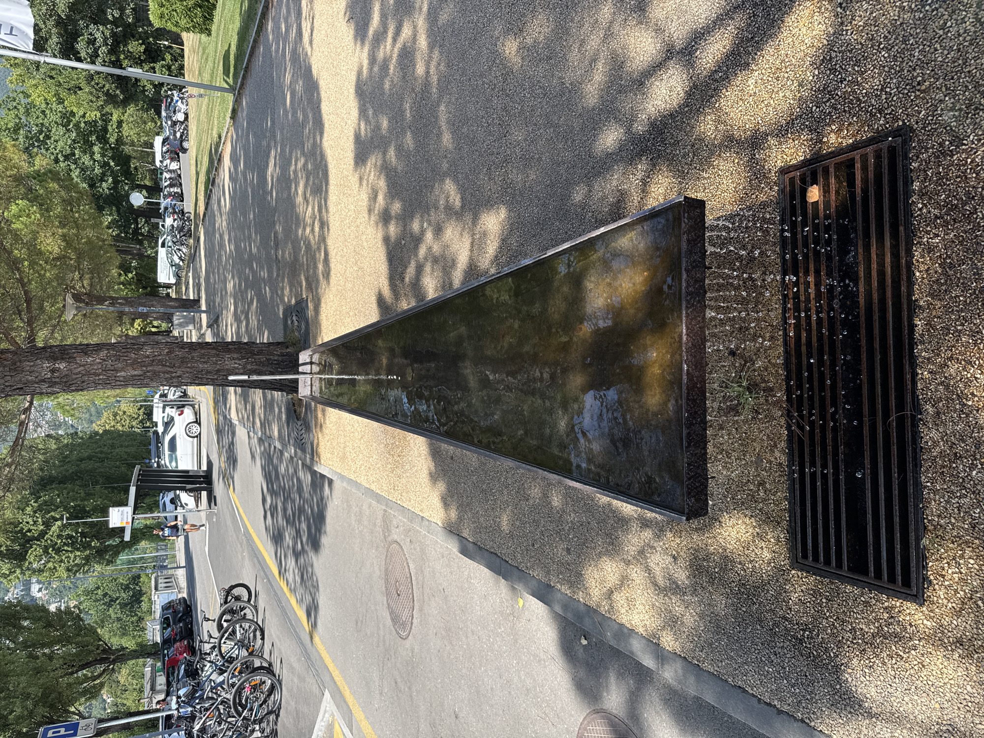 The image depicts a contemporary public water feature in the city of Locarno, Switzerland, likely situated near a park or recreational area. The water feature consists of a long, narrow trough filled with water, elevated above ground level. Water cascades from one end of the trough, flowing down onto a dark, rectangular metal grate below. The surrounding ground is covered with small, light-colored gravel. To the left, a paved pathway is visible, separated from the gravel area by a curb. On the pathway, a bicycle rack with several parked bicycles is present, and further back, a bus stop shelter is visible with a person walking towards it. In the background, cars are parked, and a line of trees runs along the edge of the paved area. The scene is bathed in sunlight, casting distinct shadows from the trees onto the ground.