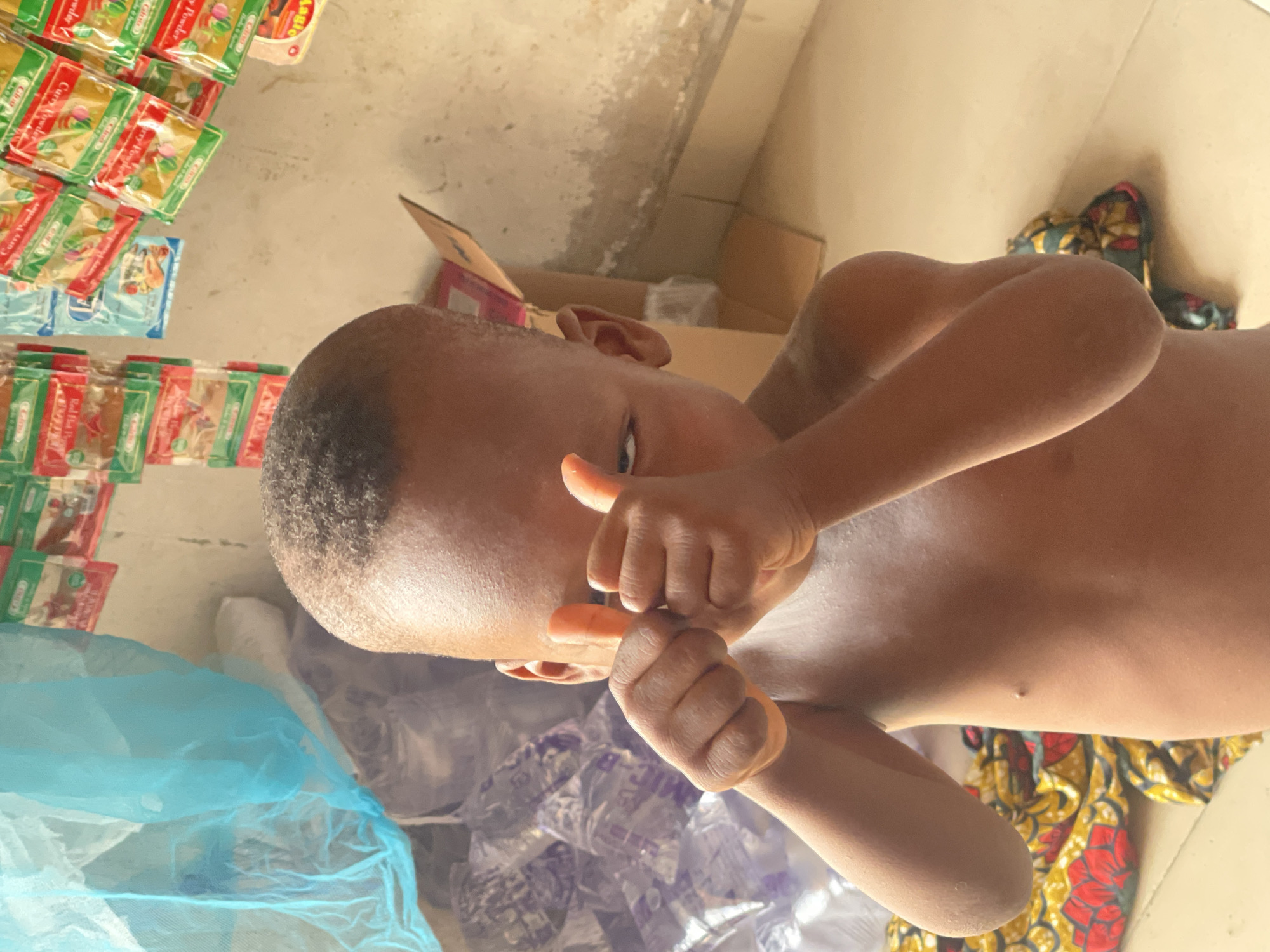 An overhead photograph, rotated 90 degrees clockwise, captures a young, dark-skinned child in what appears to be an indoor setting, likely a small shop or storage area in Lokoja, Nigeria. The child, possibly male, is shirtless and has short, dark, closely-cropped hair. He looks directly at the camera with a neutral to slightly curious expression, holding his hands up towards his face, with one thumb visible pointing upwards.
The background to the left features numerous small plastic pouches of spices or seasonings, stacked and displayed. Several green and red packages are clearly labeled "Curry Powder" and bear the brand name "Chapp". A blue-packaged item is also visible, along with another package that has the text "AGBO". To the right of the child, cardboard boxes are visible, suggesting a storage or retail environment.
The floor beneath the child features various textiles and items. A translucent blue mesh fabric, possibly a mosquito net, is visible in the lower left. Adjacent to it is a colorful patterned fabric with red, yellow, and green designs, characteristic of West African textiles like Ankara. Several clear plastic bags, appearing to contain darker, unidentifiable items, are also scattered on the floor. The overall impression is a candid moment captured indoors, potentially within a local market stall or home setting.