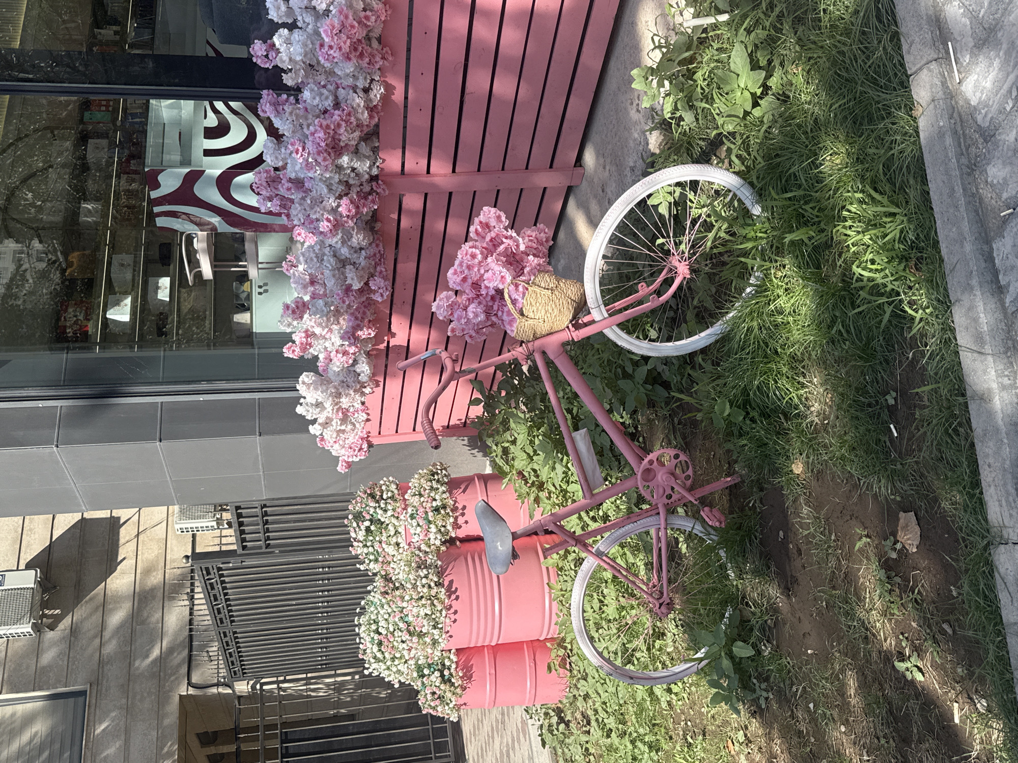 The image shows a pink bicycle with a basket filled with pink flowers parked on grass in front of a pink wooden planter box overflowing with pink and white flowers. Behind the planter is a building with large windows reflecting items inside. To the left of the planter is a black metal fence, and behind that are three large pink cylindrical planters filled with smaller pink and white flowers. Two air conditioning units are visible on the building's exterior. The scene appears to be an outdoor display outside a shop or establishment in a city. The specific location in Bishkek City, Kyrgyzstan, cannot be determined from the image alone.
