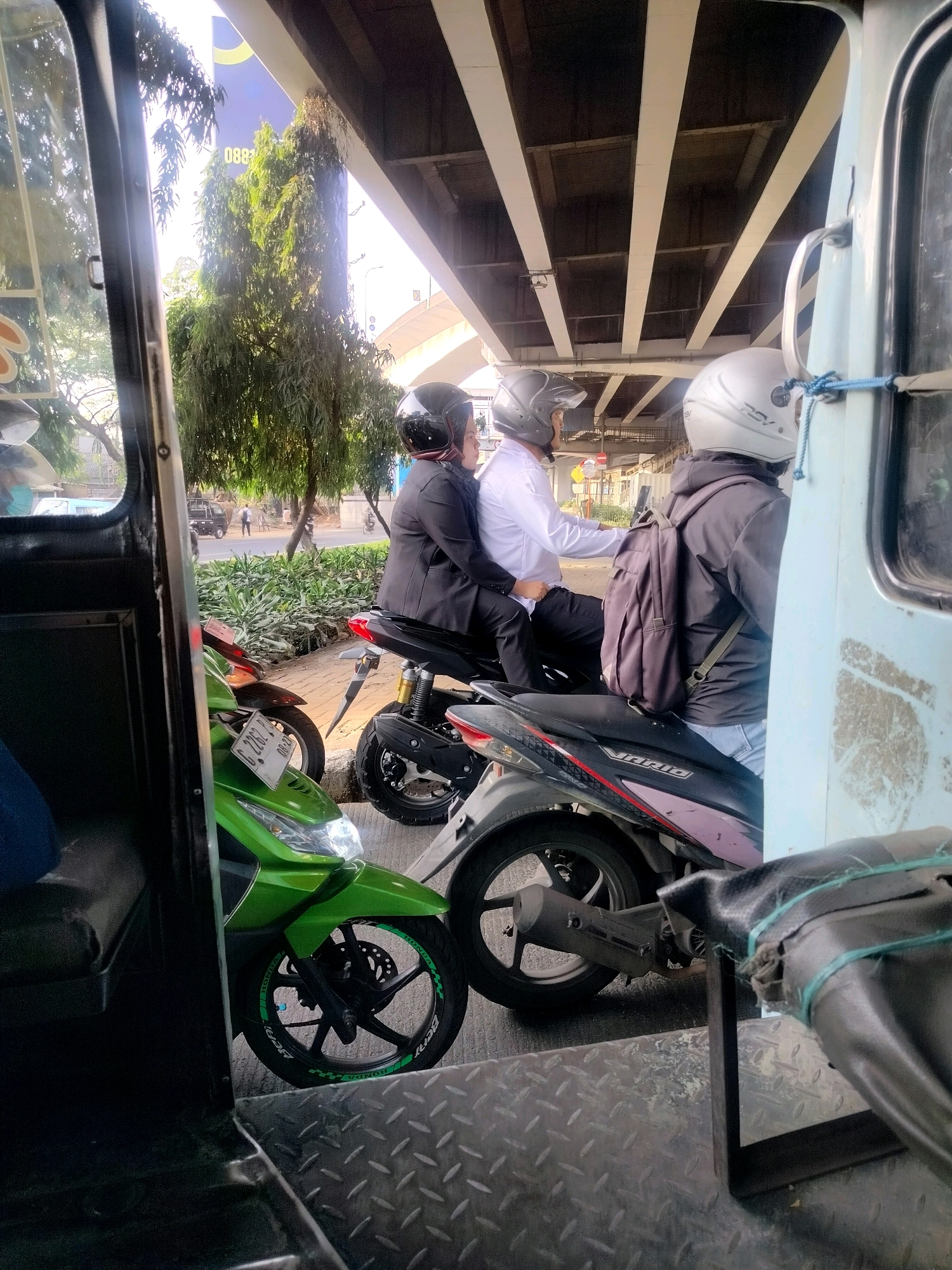 A side-view, eye-level shot captures two people on a motorcycle riding under an elevated roadway in East Jakarta, Indonesia. The motorcycle is a black Honda Vario 160. The rider, a male, wears a white long-sleeved shirt and a black helmet. The passenger, a female, wears a black jacket and helmet. She has her arms wrapped around the rider. The motorcycle is parked in front of another green motorcycle. In the background, a tall tree with lush green foliage is visible. The sky is bright and sunny.
