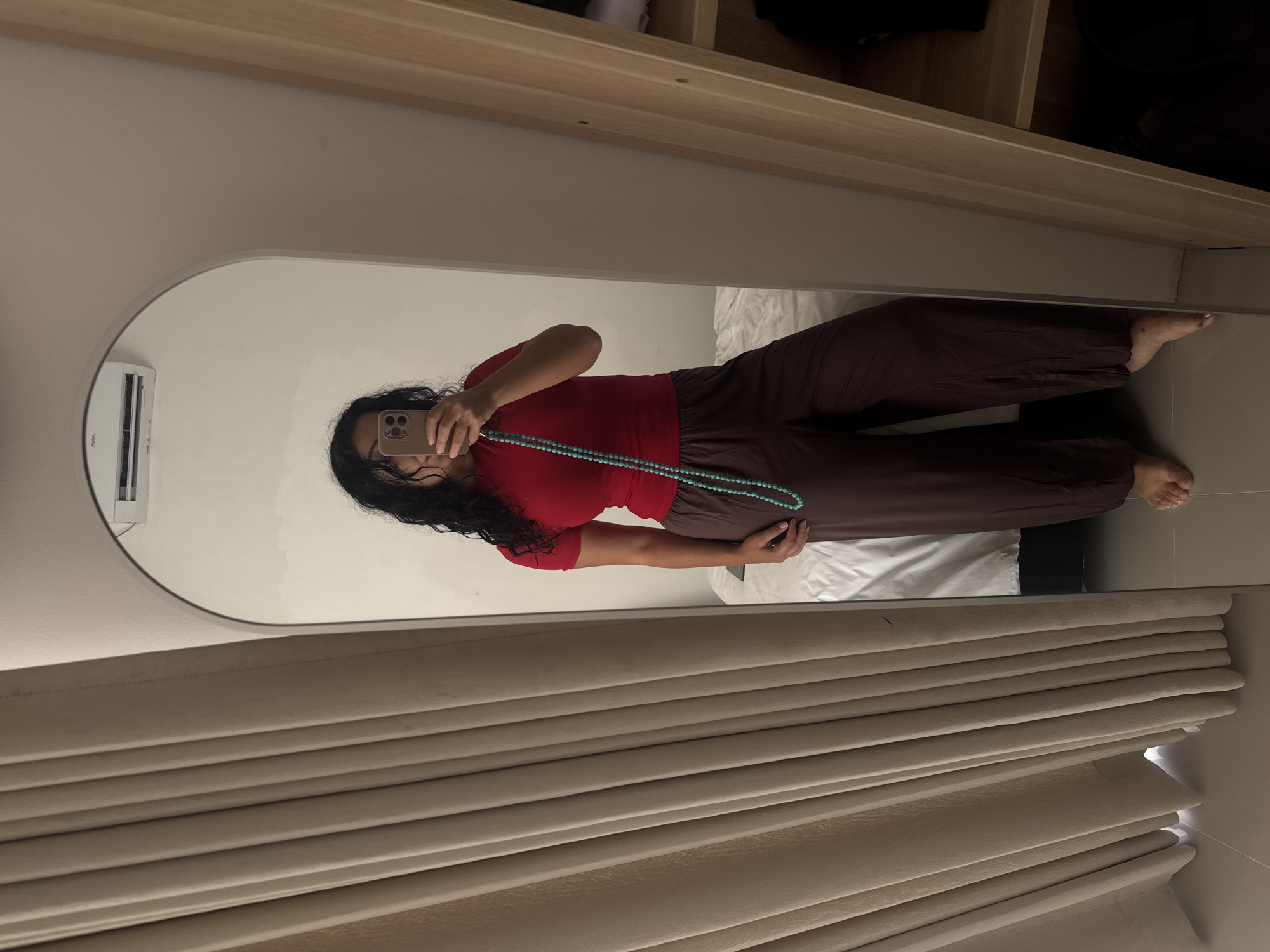 A person is reflected in a rounded-top mirror. The person is wearing a red shirt and brown pants, and holding a phone in front of their face. The person is also holding a blue beaded necklace. The mirror is in a room with beige curtains, a white air conditioner, and wooden shelving above. The room appears to be in a residence. No specific elements definitively place the image in Kuta Selatan, Indonesia.