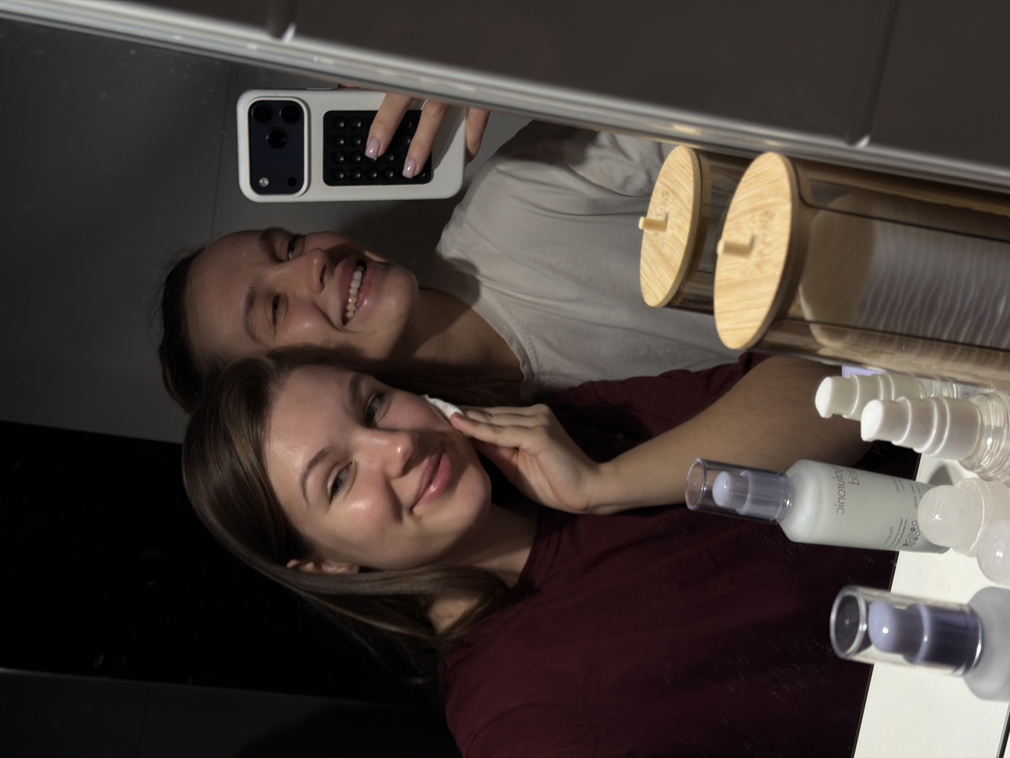 Two women are taking a selfie in a bathroom mirror. The woman in the foreground is wiping her face with a cotton pad. The other woman is behind her, holding a phone with a white and black case to take the photo. There are toiletries and containers with bamboo lids on the right side of the frame. The location is Vozdovac Urban Municipality, Serbia.