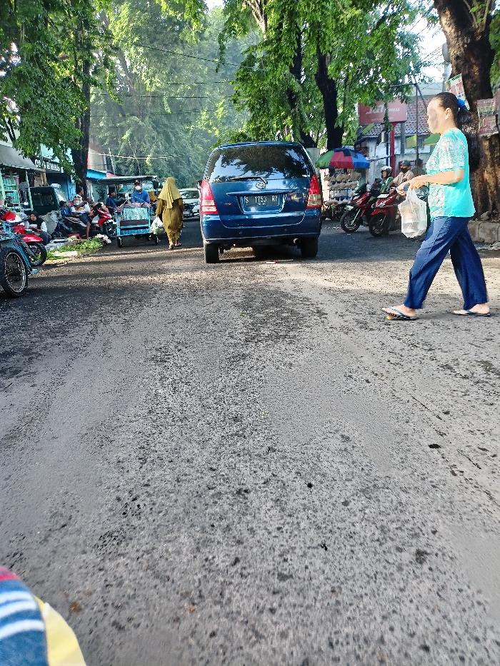 This image captures a vibrant street scene in Mojosari, Indonesia, likely during the morning or late afternoon hours given the bright, sunny lighting and clear sky. The setting is a bustling road, possibly near a market area, lined with lush green trees and informal stalls.
In the foreground, the asphalt road shows signs of wear. On the far left, a person's leg and foot, clad in colorful striped pants, are partially visible, suggesting the perspective of someone standing or sitting low. Adjacent to this, part of a blue bicycle is seen.
The midground is dominated by a dark blue Toyota Kijang Innova SUV, with the Indonesian license plate "S 1753 RY", facing away from the camera. To the left of the SUV, a woman wearing a light brown or olive green hijab and long dress walks away, carrying a small item. Behind her, a group of men are gathered around what appears to be street vendor stalls, with some individuals wearing face masks. A blue-sided pushcart is also visible among them. Several motorcycles are parked or standing along this side of the street, which features informal shops with awnings. A green awning has partial text, possibly "CLASS" or "GLAS".
On the right side of the road, closer to the camera, another woman walks away. She has dark hair pulled back and is dressed in a light blue patterned top, dark blue pants, and flip-flops, carrying a white plastic bag. Behind her, more motorcycles are parked, and another stall with a colorful umbrella displays items, potentially shoes. In the background right, a red sign with "EXPRESS" is partially visible.
The overall activity depicts daily life, with people walking, congregating, and engaging in commerce along a busy street. The presence of trees suggests an established area, and the style of the stalls and vehicles are characteristic of the region.