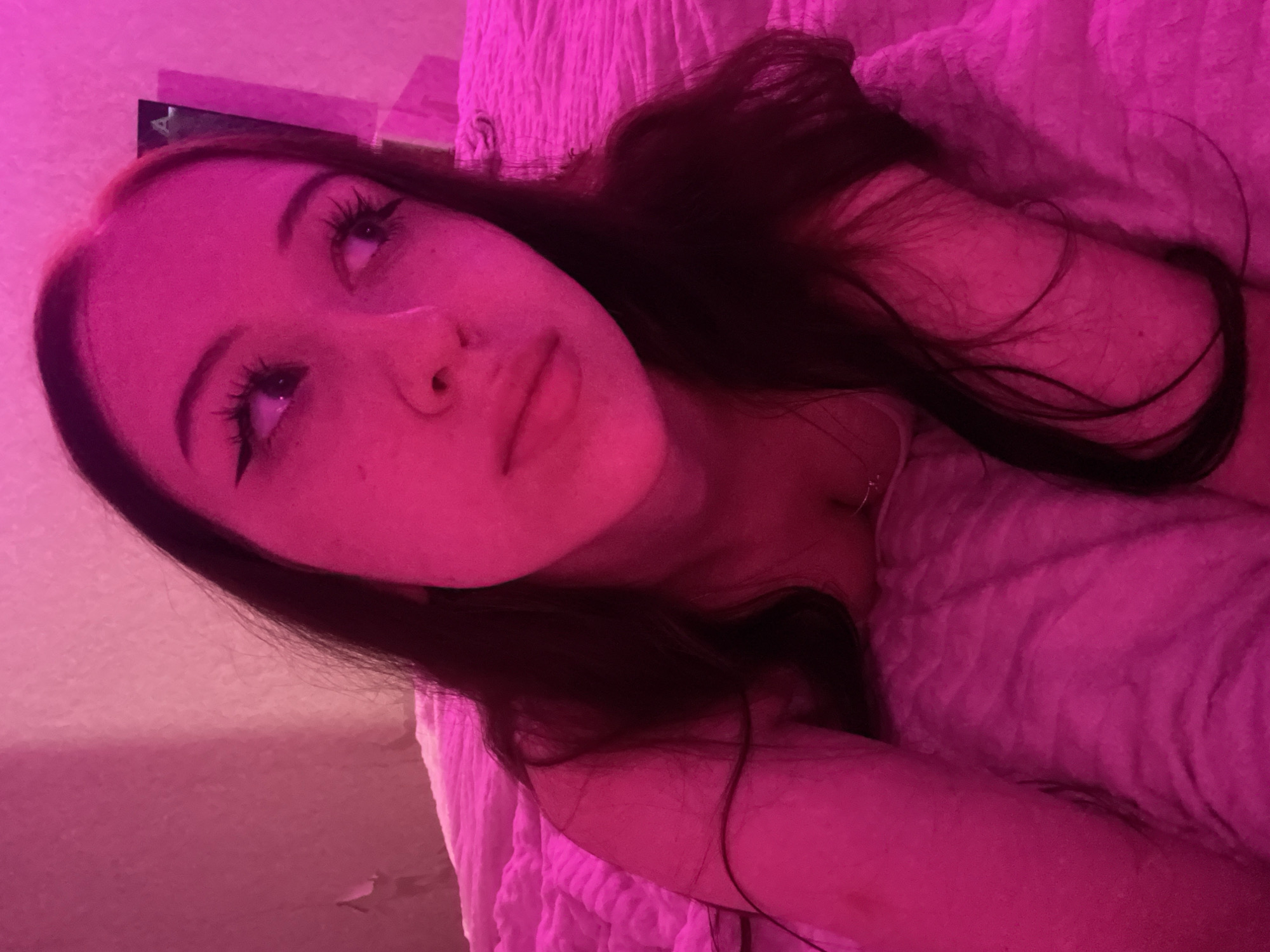 A young woman with long, dark hair and visible makeup, including eyeliner and false eyelashes, is lying on a pink, textured surface, possibly a bed or couch. The scene is illuminated by pink lighting, casting a strong magenta hue over everything. The woman is looking off to her right, with her head resting on her arm. Her expression appears contemplative or perhaps slightly melancholic. The background is a plain, textured wall, also illuminated in pink. There are no discernable elements within the image that would allow for the identification of a specific location in Votkinsk, Russia, or any other city. The image is a close-up shot, focusing primarily on the woman's face and upper body, with the surrounding environment providing context but no specific geographical clues.