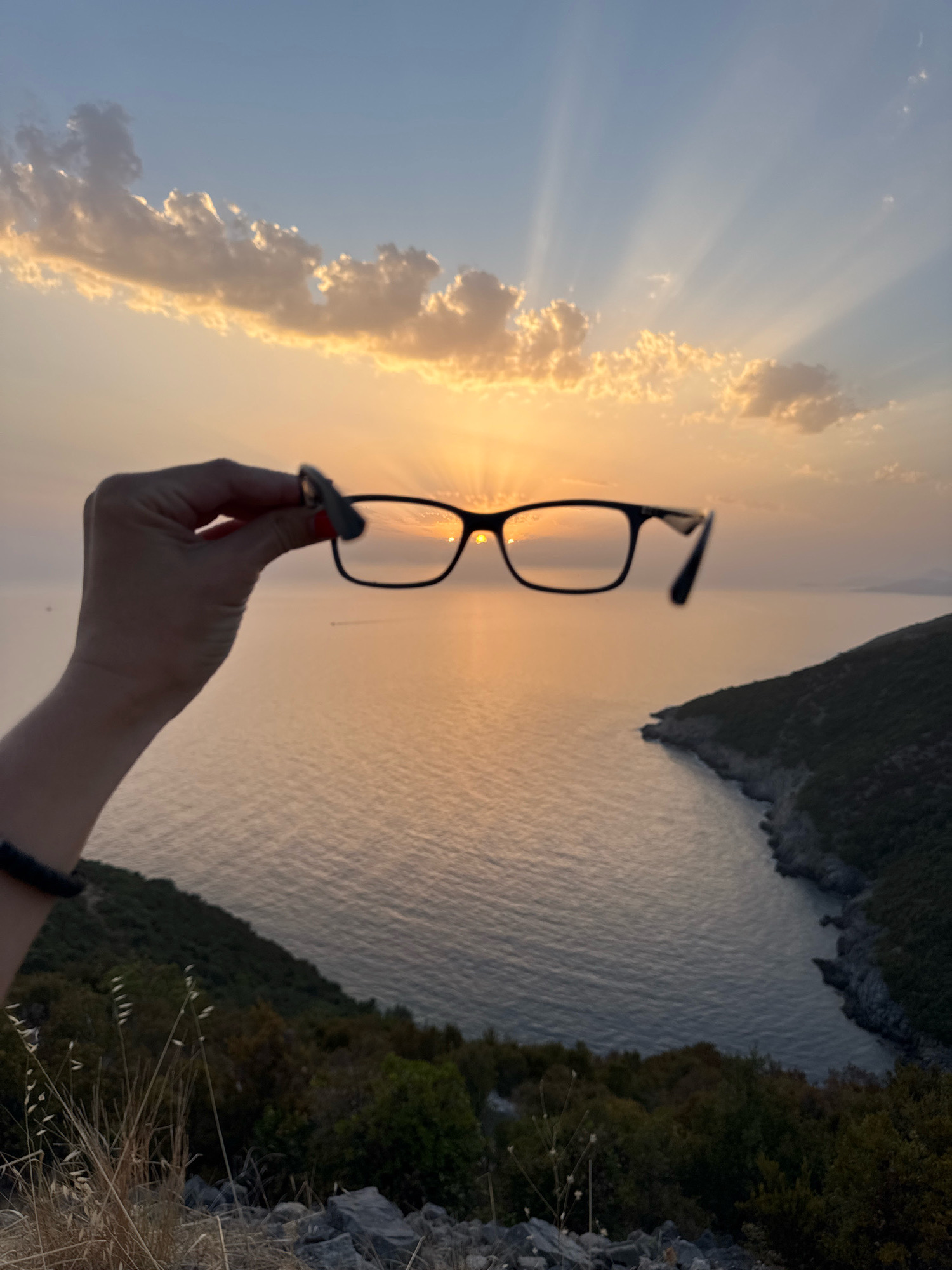 A person's hand, with red nail polish, holds a pair of black-rimmed glasses in the foreground. The glasses are positioned to frame the setting sun, which casts golden rays through the clouds and reflects on the calm sea. In the background, a lush green hillside slopes down to the water, with rocky outcrops along the coastline. The scene is set in Zagora, Montenegro, during sunset.