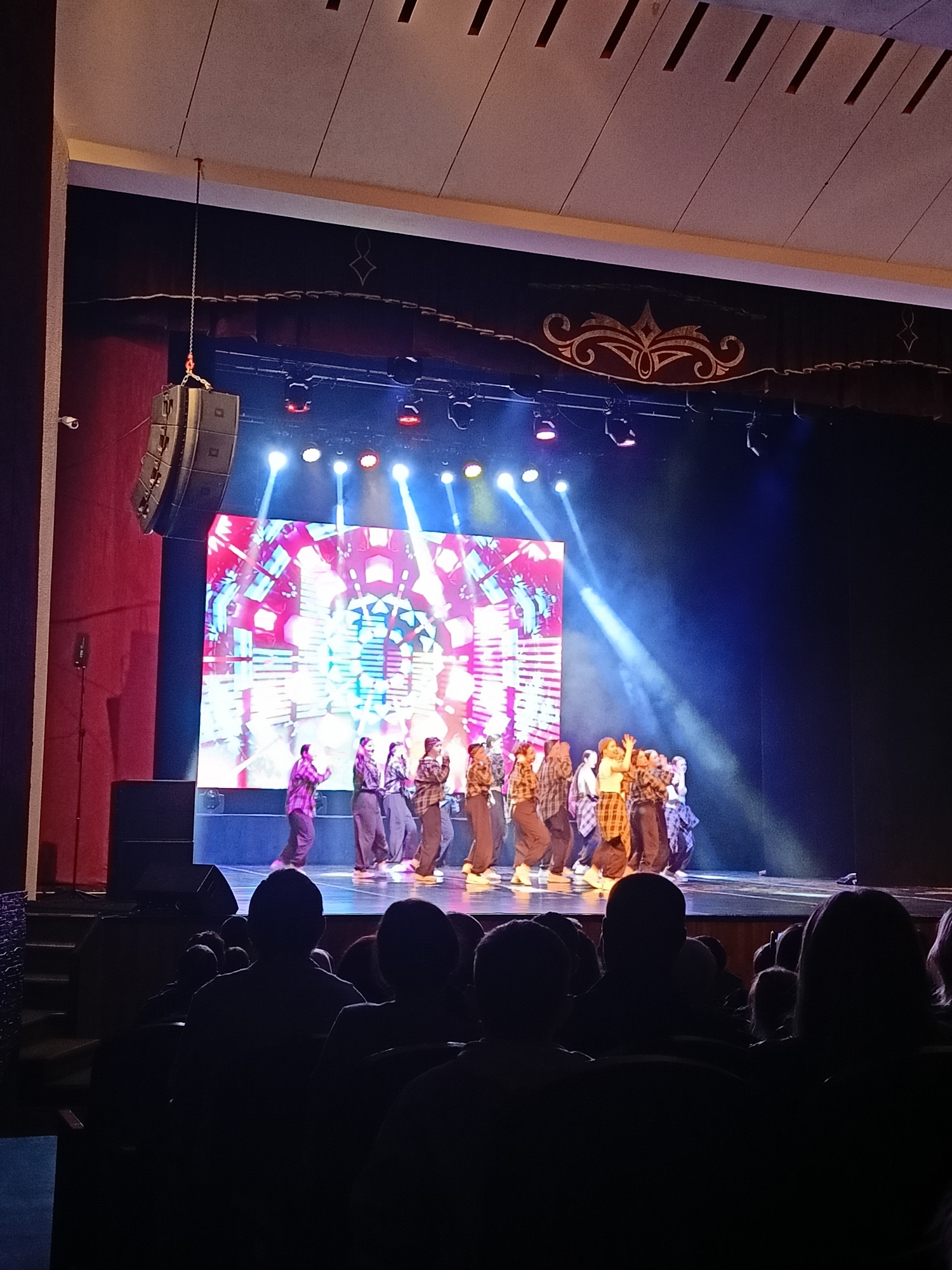 A stage performance is taking place in a theater in Chaikovsky, Russia. A group of people are dancing on a stage that is illuminated with bright spotlights and a large LED screen displaying colorful patterns. The dancers are wearing casual clothing, including plaid shirts and pants. The audience is seated in the foreground, their silhouettes visible in the dim light. A large speaker is suspended from the ceiling on the left side of the stage. Curtains flank the stage, and ornate decorative elements are visible above the stage opening.