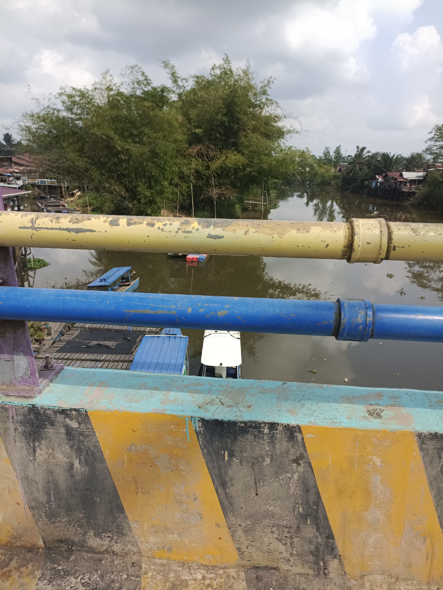 This image captures a view from a bridge in Sangatta, Indonesia. In the foreground, the weathered concrete structure of the bridge is visible, adorned with diagonal yellow and black hazard stripes. Two large pipes, one painted yellow and the other blue, span horizontally across the frame, obscuring a portion of the scene below.

Beneath the bridge, a muddy river flows, reflecting the overcast sky. Several boats are present on the water. Most notably, a blue-roofed boat with a small cabin and a white structure on its stern is positioned in the middle ground. To its left, a lighter blue boat with a canvas covering is moored. In the background, the lush green foliage of bamboo trees and other vegetation lines the riverbanks. Further in the distance, a cluster of houses with red roofs is visible, suggesting a small settlement along the river. The overall atmosphere is one of a humid, tropical environment, likely during the day given the ambient light. There are no people visible in the frame.