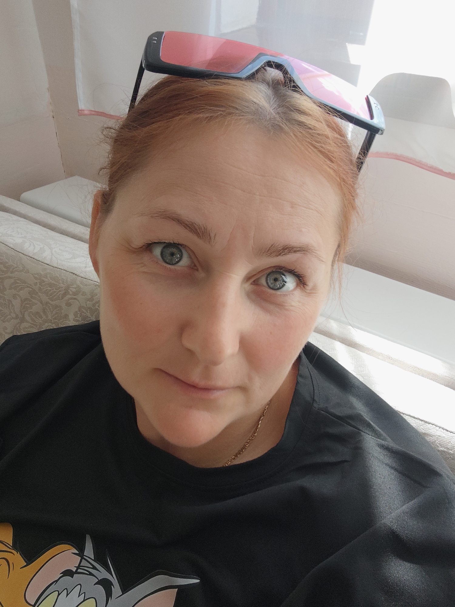 The image shows a close-up, selfie-style shot of a woman with red hair, wearing a black t-shirt with a cartoon print of Tom and Jerry. She has pink-tinted sunglasses perched on her head. She is looking directly at the camera with a neutral expression. The background appears to be an indoor setting with a textured couch and sunlight streaming in from a window or skylight, casting shadows. The image does not contain any information to indicate its location in the city of Марковский, Russia.