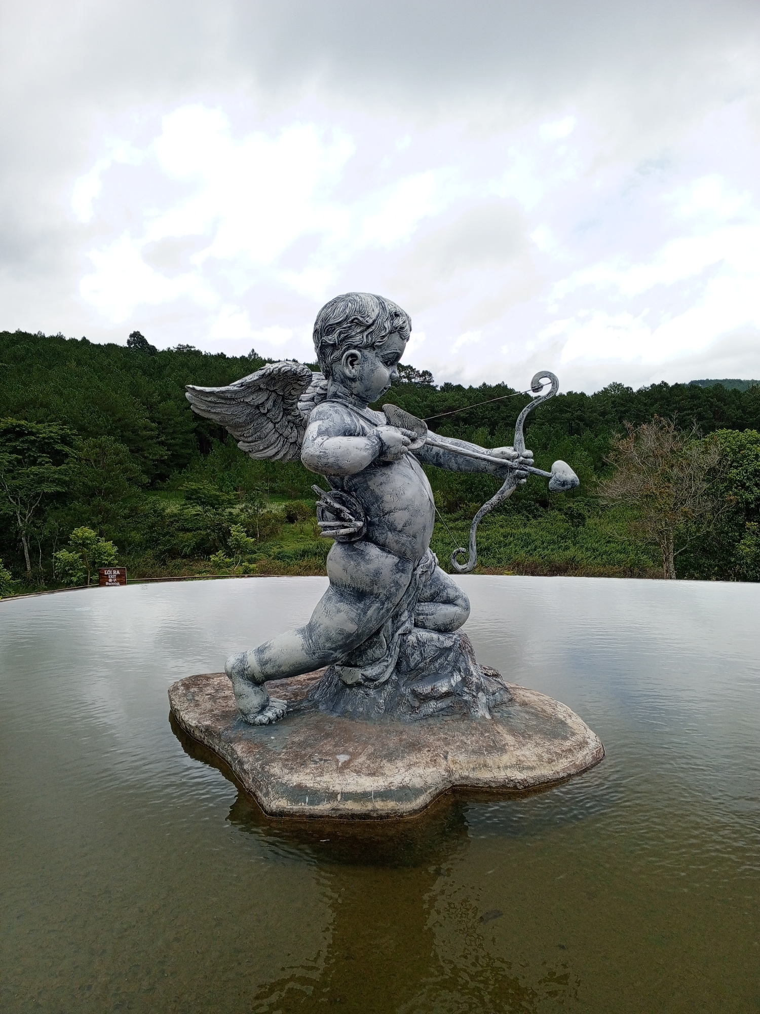 A stone sculpture of Cupid, a winged cherub drawing a bow and arrow, stands on a rock in a body of water. The sculpture is grey and weathered, with intricate details on the wings and the cherub's body. The cherub is depicted in a dynamic pose, aiming the arrow forward. The water around the rock is calm, with ripples reflecting the cloudy sky. In the background, a lush green forest covers rolling hills. A wooden sign with text is visible near the edge of the water. The overall scene suggests an outdoor garden or park setting. This statue is located at the Tuyen Lam Lake in Da Lat, Vietnam.