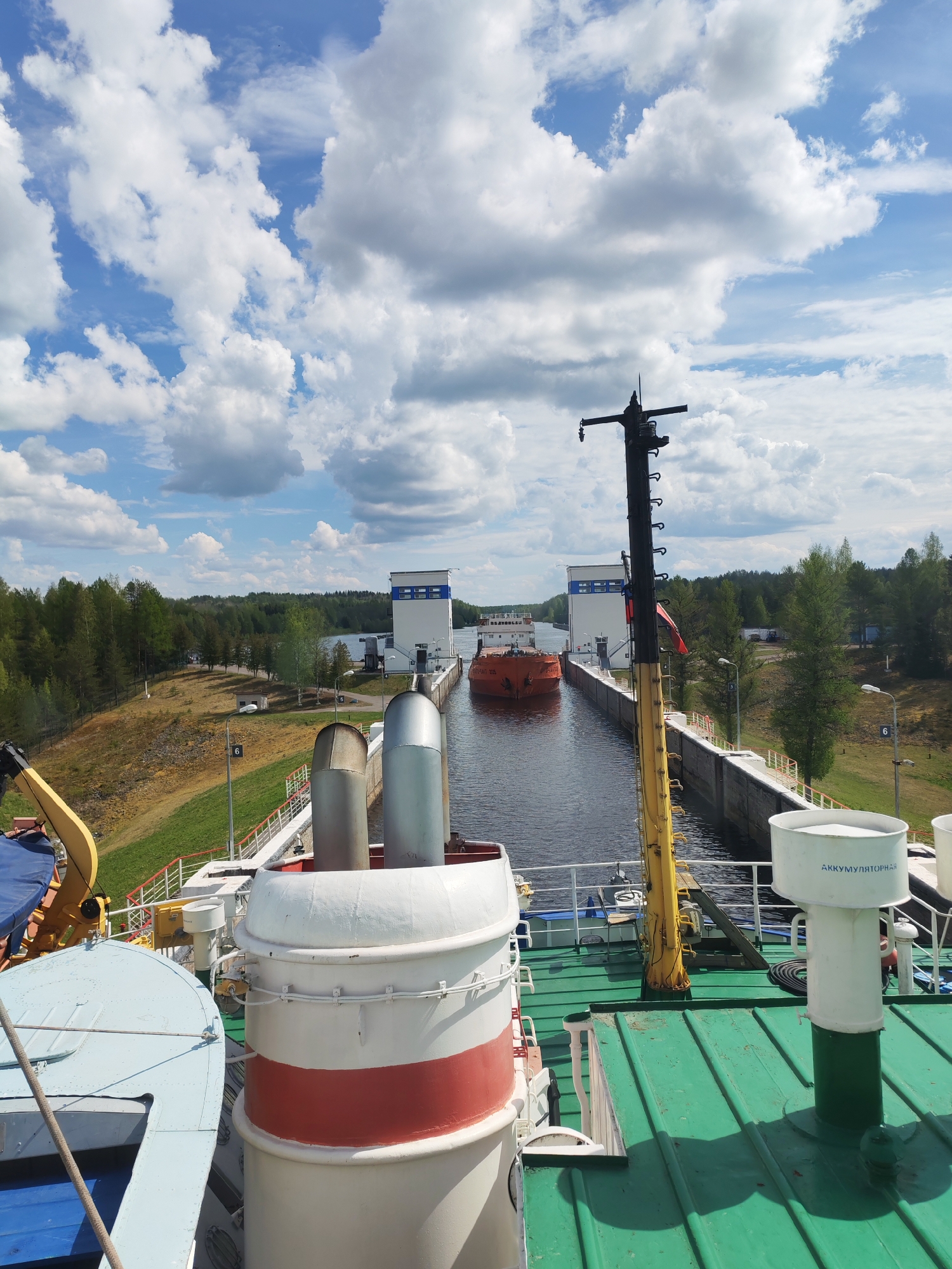 The image presents a view from the deck of a vessel, likely a ship or a large boat, situated within the Volga–Baltic Waterway near Devyatiny, Russia. The waterway is characterized by a series of canal locks, and the foreground of the image is dominated by the ship's infrastructure. The vessel is approaching a lock system.
The lock is flanked by white structures, possibly control towers or service buildings, on either side. A large orange cargo ship or barge is already inside the lock. On the deck of the ship is a mast, painted black and yellow, along with various ship components.
The surrounding environment includes forested areas, open grass slopes, and a bright sky with scattered clouds. The scene is captured in daylight, suggesting clear weather conditions.