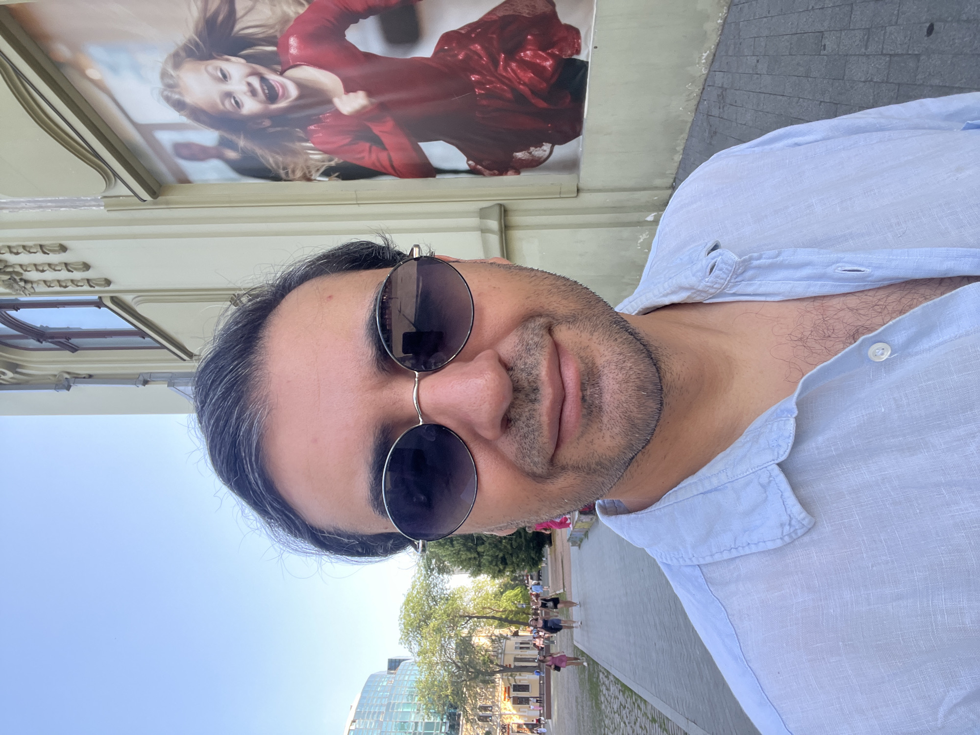 A man is taking a selfie outdoors in a city. He is wearing a light blue linen shirt and sunglasses. In the background, there is a large advertisement for a young girl in a red dress. The man's face is partially visible, and he appears to be smiling. The scene suggests it is daytime, with a clear sky and sunlight. The location appears to be a pedestrian walkway or street lined with buildings and trees. People can be seen walking in the background, indicating a public area. The overall mood seems casual and relaxed.