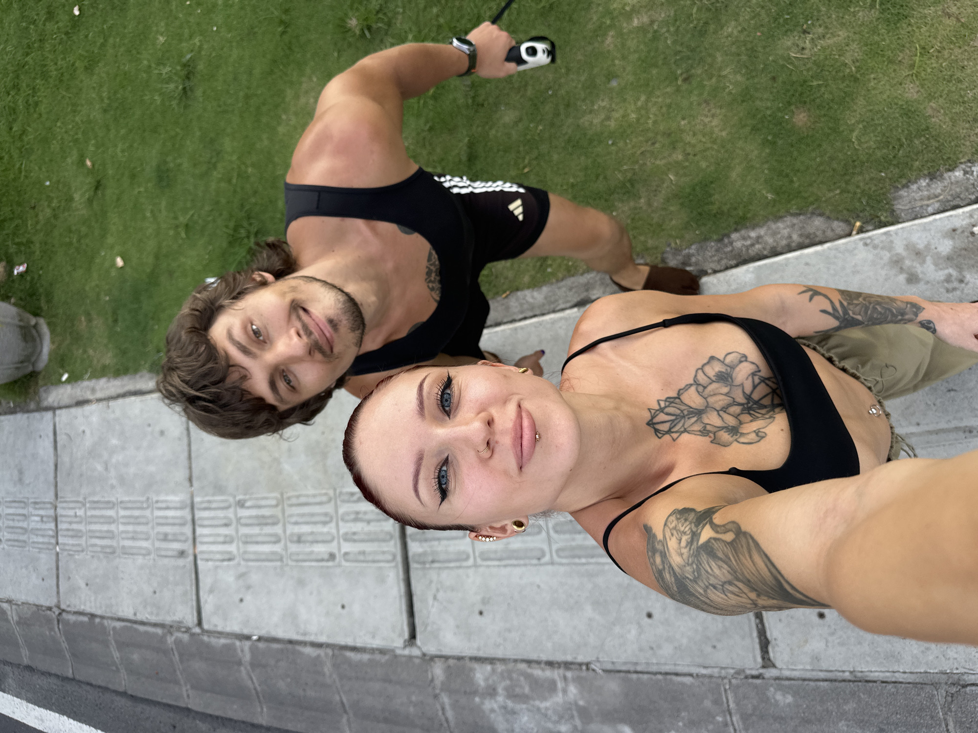Two individuals are positioned at the edge of a concrete pathway in Dalung, Indonesia. The pathway is characterized by a tactile paving surface intended for pedestrian navigation, and it is bordered by a strip of grass. A male is situated to the left of the frame; he has brown hair, a light beard, and is wearing a black tank top and shorts. He is holding a white and black object, possibly a leash handle, in his right hand. To the right, a female is taking a selfie. She has short red hair, multiple piercings, and tattoos on her chest and arm. She is wearing a black top.