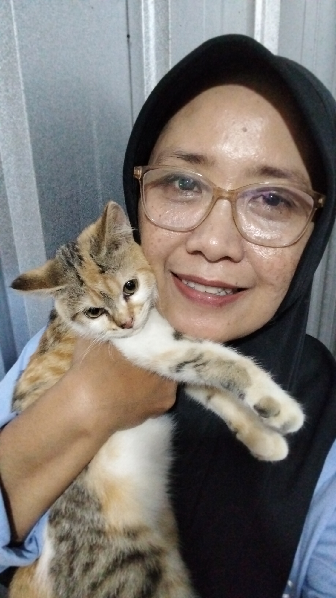 A woman wearing glasses and a black hijab is holding a calico kitten up to her face. The woman is smiling and the kitten is looking towards the viewer with a slightly concerned expression. The background appears to be an indoor setting with a textured metal wall. The lighting is dim, suggesting an indoor photo taken at night. The image is cropped closely, focusing on the woman and the kitten. The woman is wearing a light blue shirt. The kitten has patches of orange, black, and white fur. The woman's glasses are light brown. The setting does not contain any specific details that would indicate a location in Tegalombo, Indonesia.