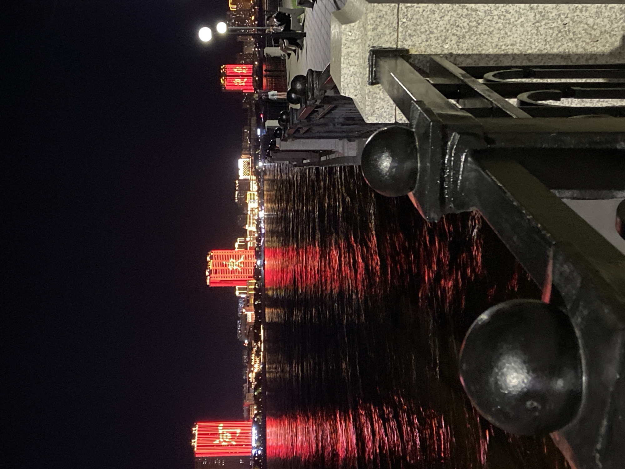 The image shows a waterfront scene at night in Blagoveshchensk, Russia. In the background, across a body of water that reflects the lights, are several illuminated buildings. Two prominent buildings on the left and one on the right are lit with bright red characters, which appear to be Chinese. Other buildings in the mid-ground are illuminated with yellow and white lights. A railing with decorative black spherical finials runs along the foreground, separating the viewer from the water. On the walkway behind the railing, several people are visible. One person stands near the railing, while others are seated on benches. Lampposts with white lights illuminate the walkway. The water is dark, with ripples catching the red reflections from the buildings. The overall atmosphere is that of a vibrant city at night.