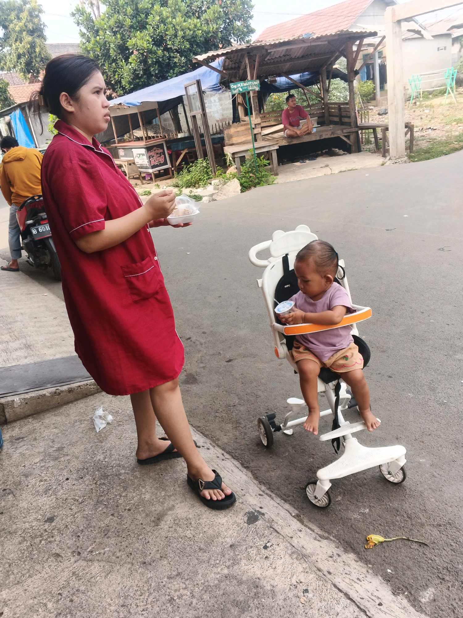 A woman in a red dress stands on a paved surface holding a container of food and a plastic bag. A toddler sits in a white stroller, holding a cup. Behind them, a man in a yellow jacket stands next to a motorcycle. In the background, a wooden shelter with a corrugated roof is visible, and a building with a tiled roof. The scene appears to be a street in Kadu, Indonesia, with a residential area and market stalls.
