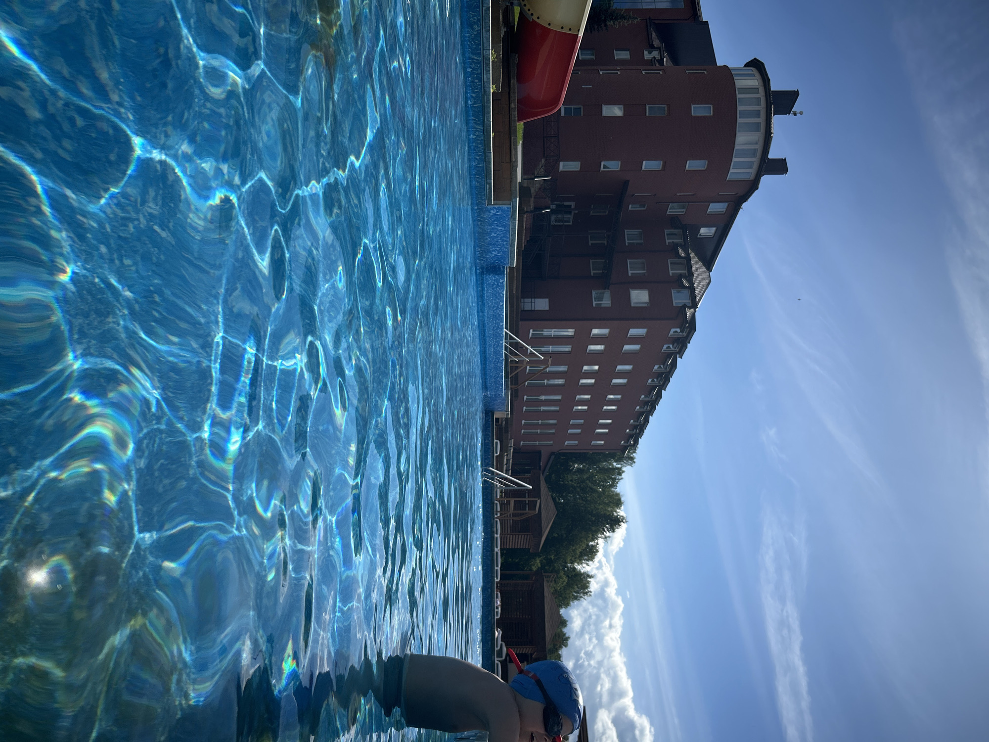 The image depicts a bright, sunny day at an outdoor swimming pool, likely in Yubileyny, Russia, given the context. In the foreground, the crystal-clear blue water of the pool dominates, with sunlight creating shimmering reflections and refractions on its surface. A young child wearing a blue swim cap and goggles is partially visible in the water on the right side of the frame, looking upwards and to the right. In the background, a multi-story brick building with numerous windows and a distinctive round turret is visible. Adjacent to the building, a red and yellow water slide curves into the pool area. Further back, several small wooden gazebos or cabanas are situated, suggesting a resort or recreational facility. The sky is a vibrant blue with wispy white clouds. The overall scene conveys a sense of leisure and enjoyment on a summer day.