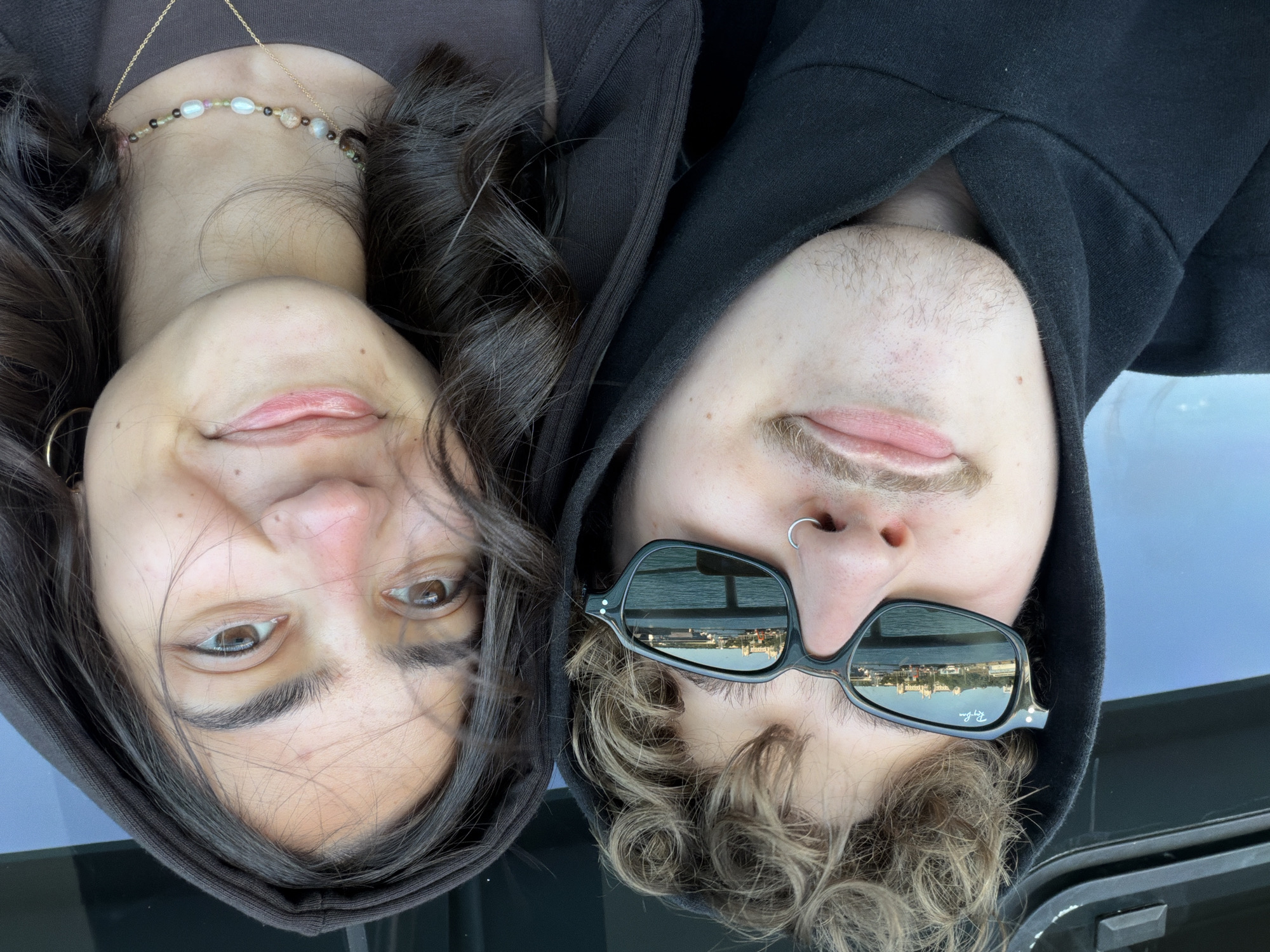 Two people, a woman and a man, are shown in a vertically oriented, close-up shot, apparently lying down. The woman is on the left wearing a brown hooded top, while the man is on the right in a black hoodie and sunglasses. The woman has long dark hair and a beaded necklace with a gold chain. The man is wearing a nose ring, has light brown hair, and his sunglasses reflect a waterfront view. The reflection shows buildings along the shoreline in what appears to be Kadıköy, Turkey. The background is a slightly blurred blue-grey surface.