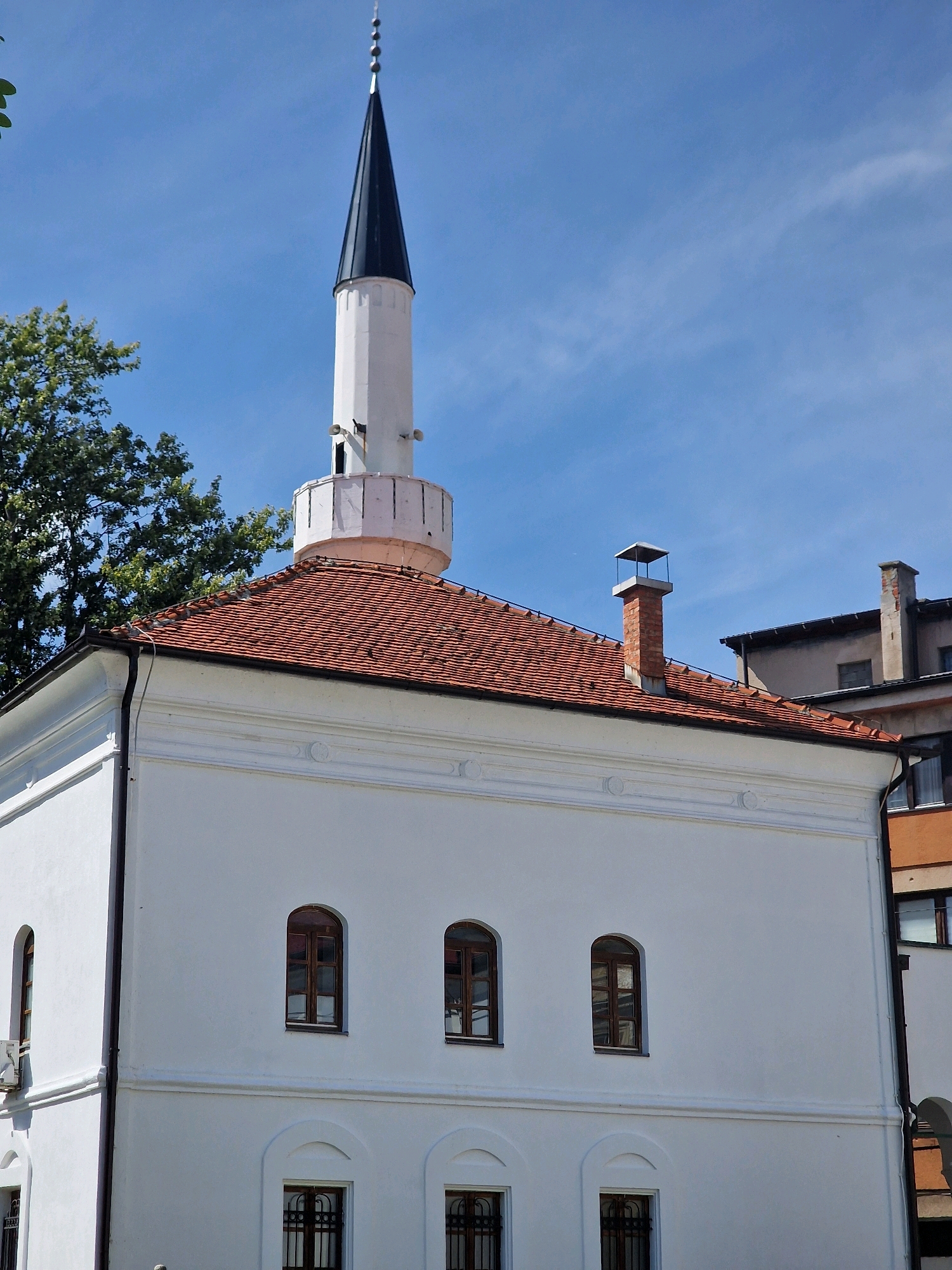 The image shows the mosque of Gazi Mehmed-paša Kukavica in Travnik, Bosnia and Herzegovina. It is a sunny day with a clear blue sky. The mosque is a white building with a red tile roof and a tall, white minaret topped with a black spire. There are several arched windows on the mosque. A chimney is visible on the roof of a neighboring building. The mosque is surrounded by trees and other buildings.