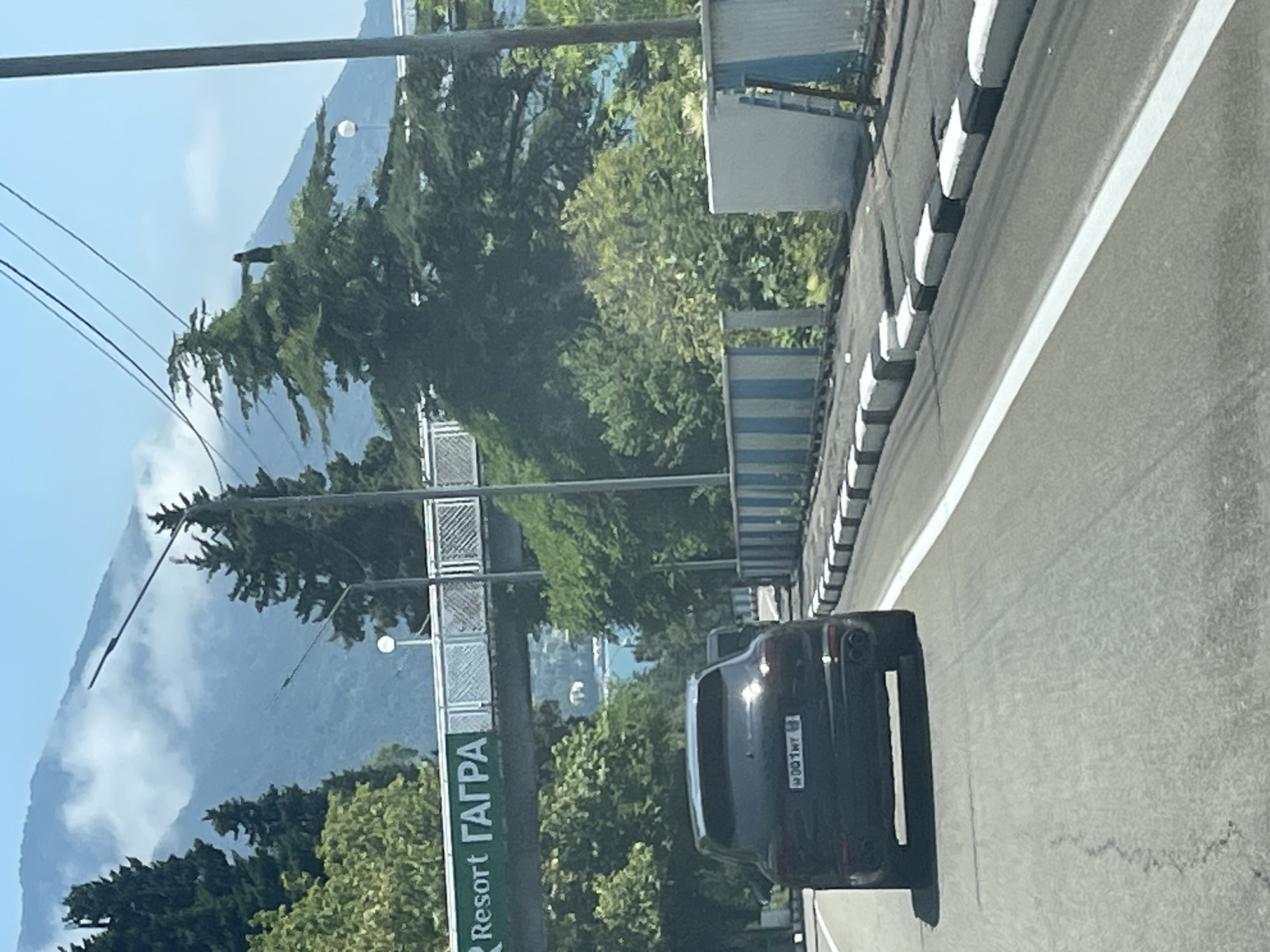 A black SUV drives on a paved road in Abaaata, Abkhazia, beneath a clear blue sky and distant, cloud-shrouded mountains. The SUV is in the foreground, facing away from the camera, with its license plate visible. A green sign with white lettering reading "Resort ГАГРА" is suspended over the road in the middle ground. Trees line the sides of the road, and a body of water is visible in the distance beyond the trees. A pedestrian overpass with a metal railing crosses the road in the upper middle ground. A striped barrier, alternating between blue and white, runs along the right side of the road.