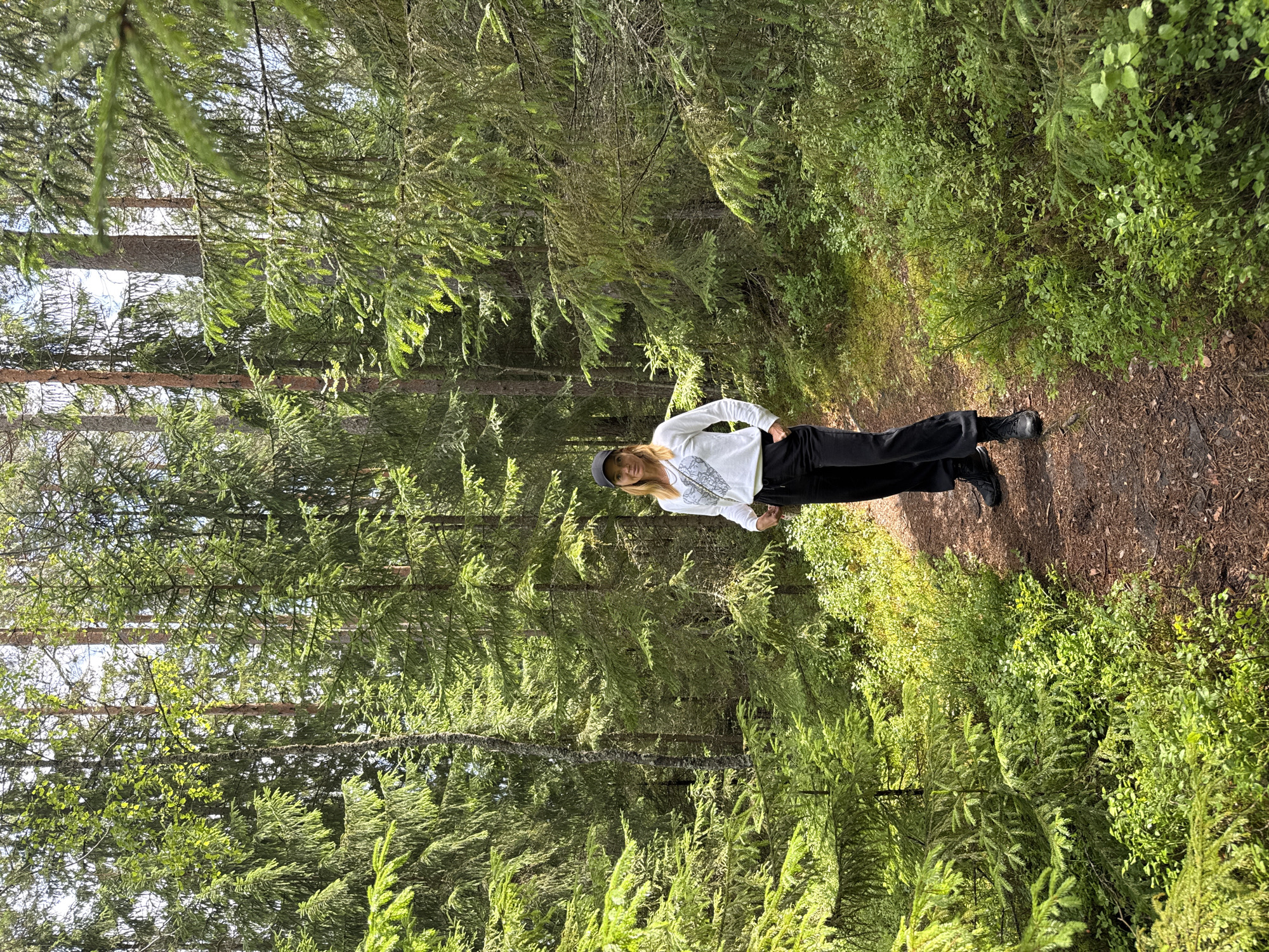 A woman stands on a dirt path in a dense coniferous forest. She is wearing a black baseball cap, a white long-sleeved shirt with a black and white pattern on the front, black pants, and black boots. The path is surrounded by green undergrowth and tall evergreen trees. The lighting suggests it is daytime. The location is described as a forest in the city of Orekhovo, Russia.