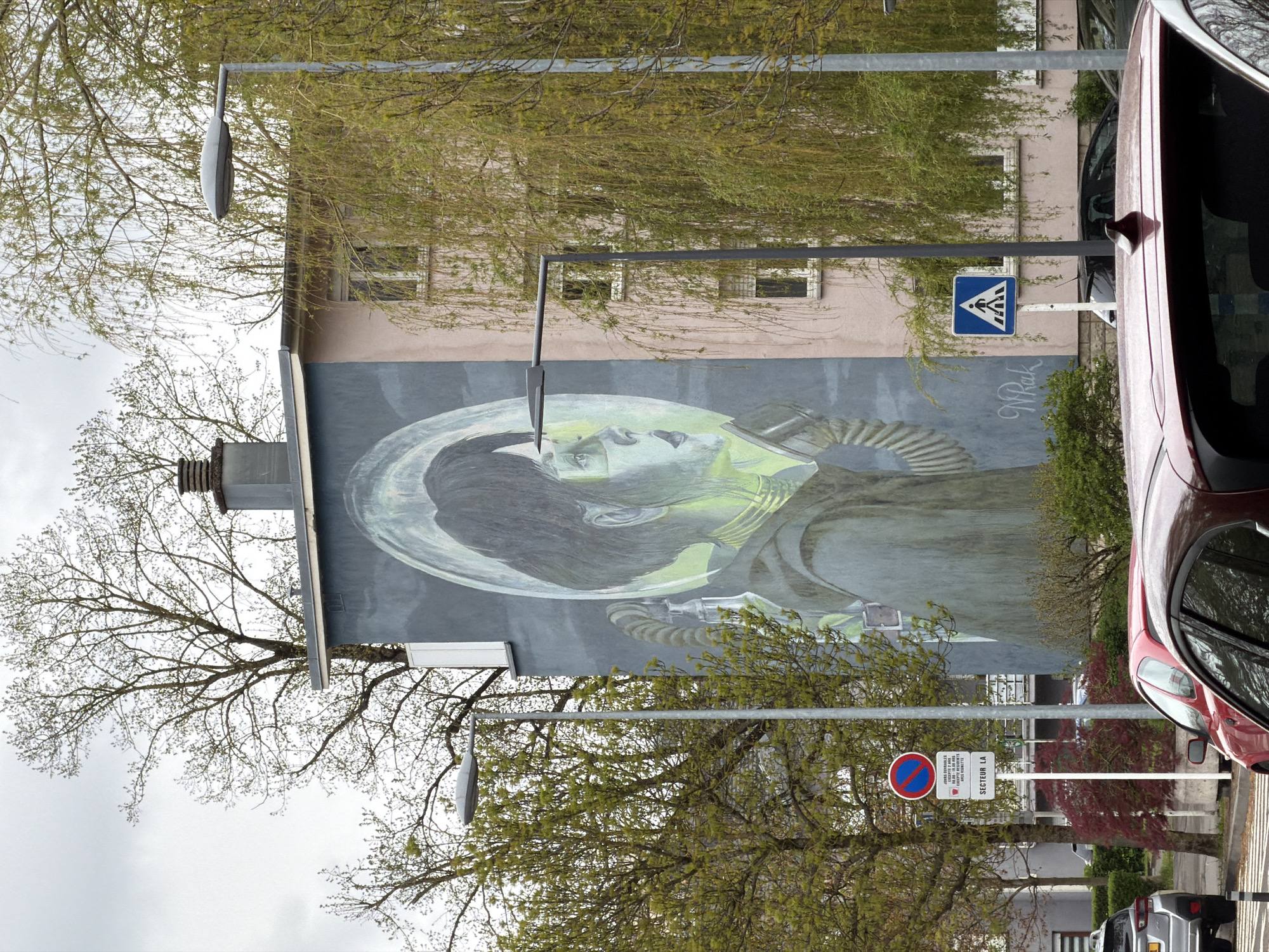 The image depicts a mural on the side of a building. The mural features a person wearing what appears to be a space helmet, depicted in shades of gray and green. The building is light-colored, possibly pink or beige, with multiple windows. A street light and trees partially obscure the view of the building and mural.
A pedestrian crossing sign is visible near the mural. Also present is a traffic sign indicating "SECTEUR LA," along with further text in French related to parking restrictions. A red car is parked close to the building. The overall setting suggests an urban environment. The location is Esch-sur-Alzette, Luxembourg, as indicated in the prompt.