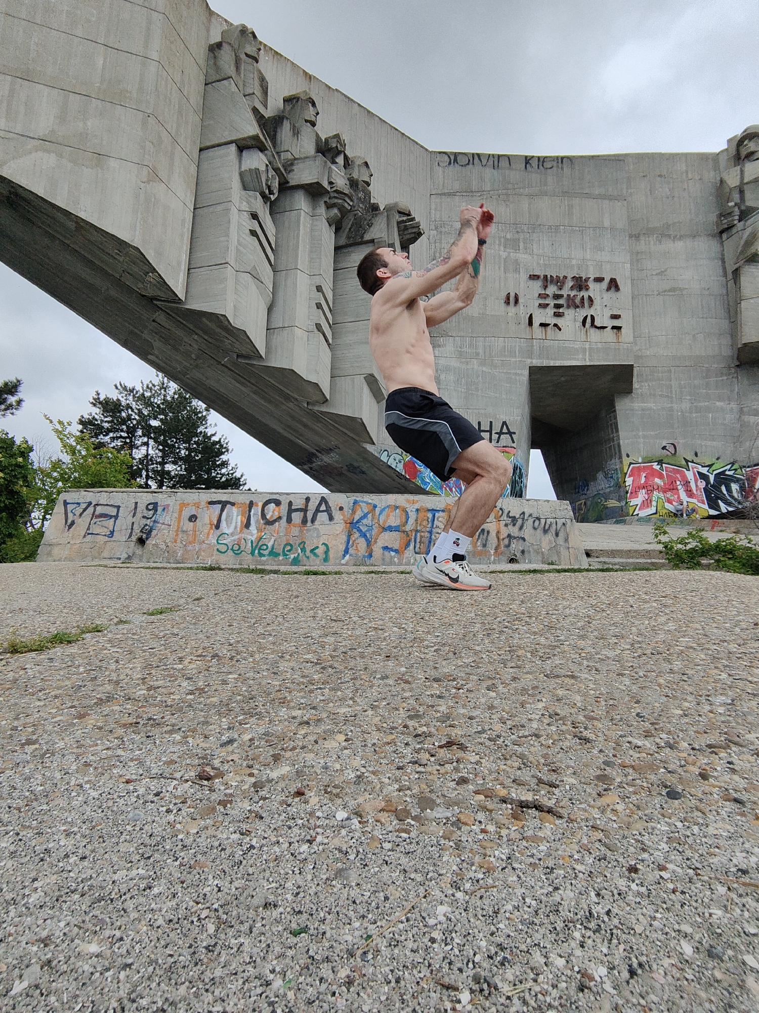 The image shows a shirtless man in athletic shorts and sneakers in the city of Varna, Bulgaria. He is captured mid-motion, likely during a workout, with his arms raised. In the background is a large, brutalist concrete structure featuring reliefs of figures. Graffiti is visible on the base of the structure. The ground is covered in a mix of gravel and concrete. Trees and greenery are visible in the mid-ground behind the graffiti. The sky is overcast.