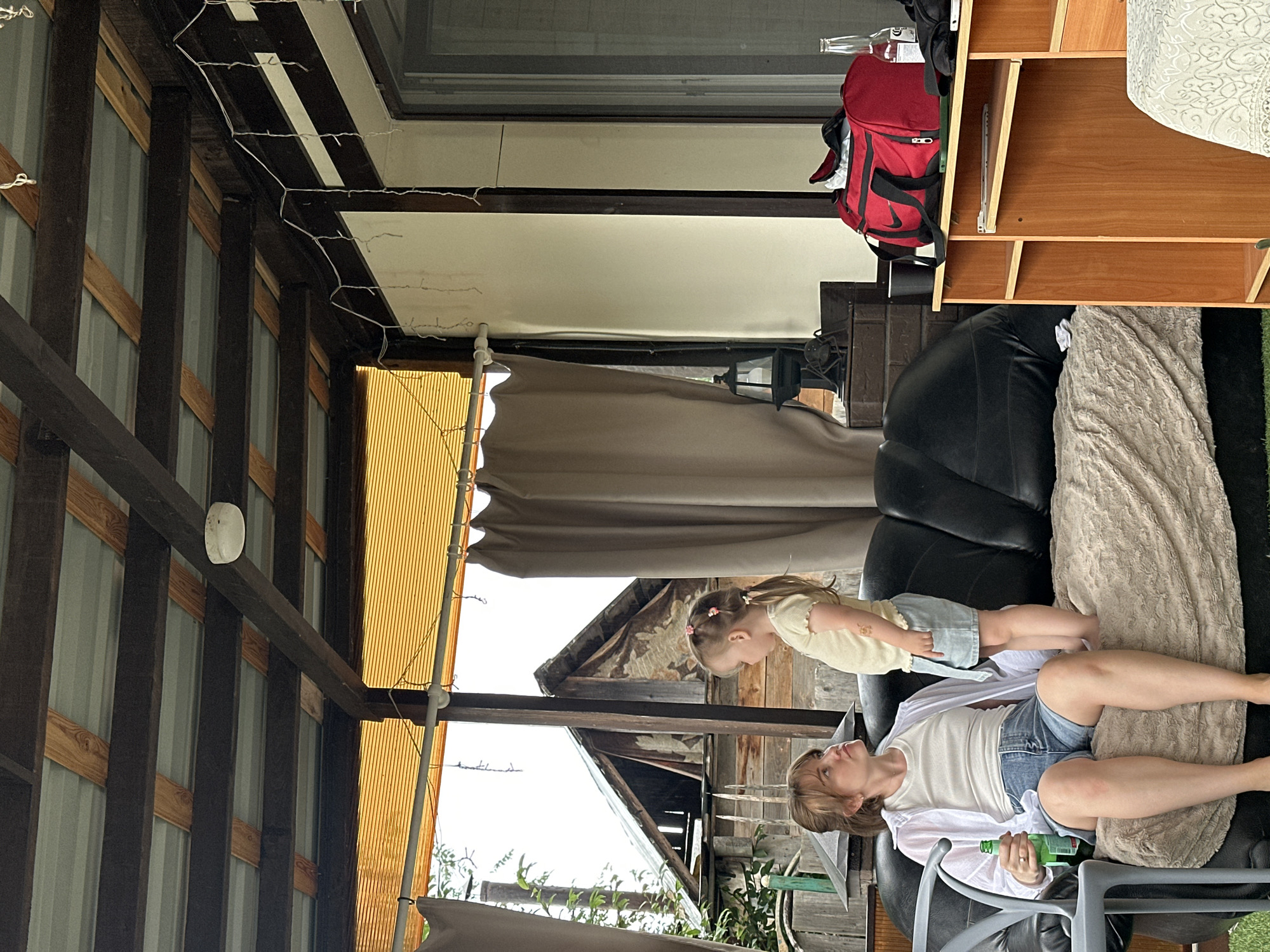 A woman and a young child are sitting on a sofa under a covered outdoor area. The woman is wearing a white button-down shirt, a white tank top, and denim shorts. She is holding a green bottle. The child is wearing a yellow top and denim shorts and standing on the sofa. Behind them is a rustic wooden structure with a corrugated metal roof. The covered area has a wooden ceiling with exposed beams and is decorated with string lights. To the right of the sofa is a wooden cabinet with a red bag and a bottle on top. The scene appears to be in a backyard or a rural setting, likely in the city of Sylva, Russia, given the architecture and vegetation.