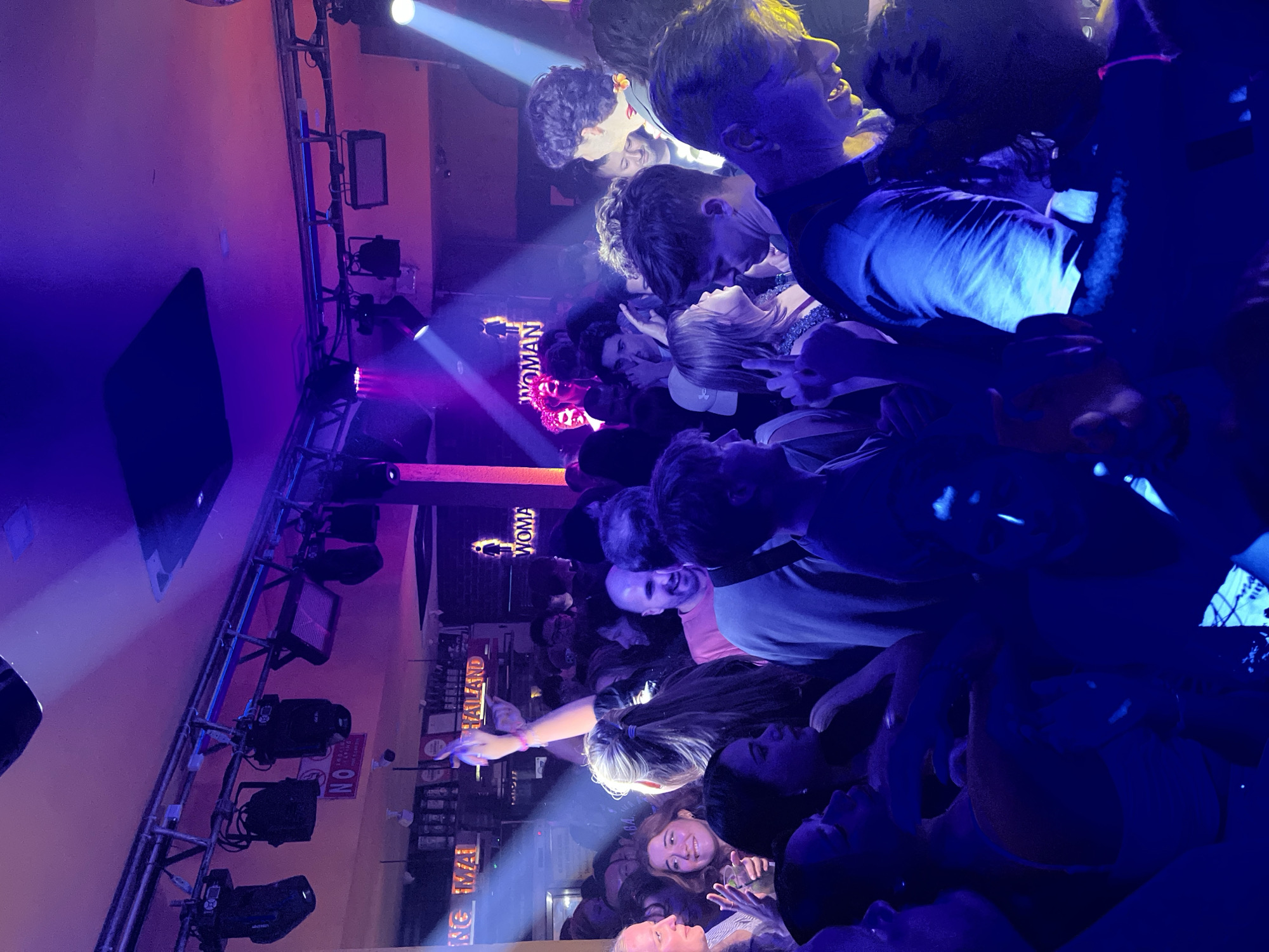 This media file captures a lively indoor scene within a crowded nightclub or music venue, taken from the perspective of someone within the audience. Numerous people, primarily young adults, are packed closely together, illuminated by a vibrant array of artificial lights.
The lighting is a dominant feature, with strong beams of blue and purple light cutting through the atmosphere, alongside bright white spotlights. This creates a dynamic and energetic ambiance. Many individuals in the crowd appear engaged and joyful; some are smiling, talking, or looking towards the front of the venue. One woman in the mid-ground is distinctly seen pointing forward.
To the left, a metal truss system holds various stage lighting equipment, including par lights, moving head fixtures, and LED panels. In the background, behind the heads of the attendees, a structure resembling a bar or decorative wall is visible.
Several pieces of text are legible within the image. Prominently, illuminated signs displaying "THAILAND" and "CHIANG MAI" in capital letters are visible, confirming the location context of Chiang Mai. Other signs include a restroom indicator marked "WOMAN," and a "NO SMOKING" sign. The equipment also shows the brand "ALOHA." The overall scene suggests an evening event filled with excitement and social interaction.