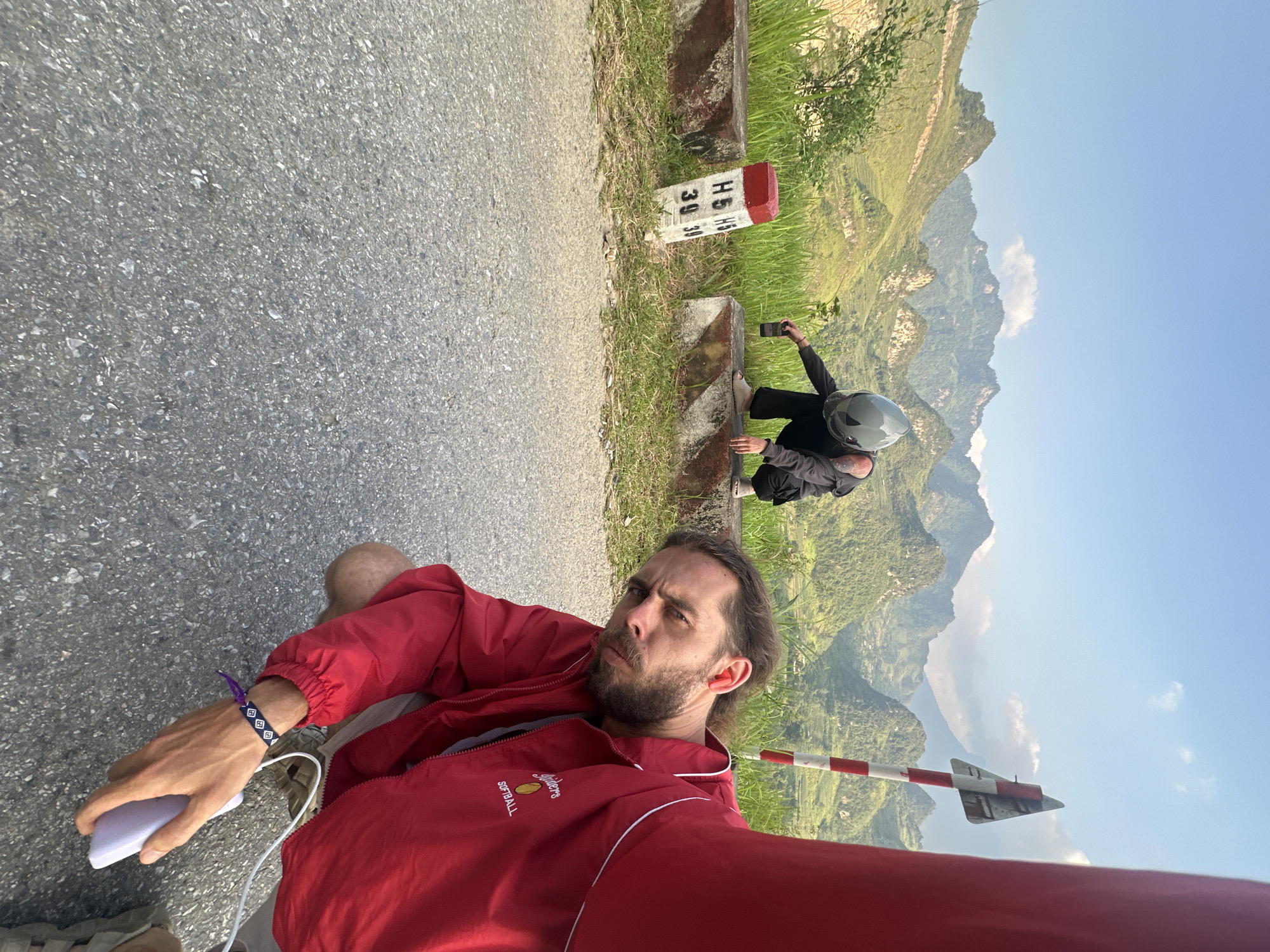 The image is a selfie taken by a man with a beard, wearing a red jacket with a white stripe, and a woven bracelet. He is sitting on an asphalt road, holding a white object, possibly a phone or a power bank. A white cable is connected to it. Another individual is visible in the background, kneeling and holding a phone, near a red and white kilometer marker with the text "H5" and "39" "38" "30". The background features a mountainous landscape with green vegetation, likely taken in Du Già Commune, Vietnam. A red and white pole is also visible in the foreground, near the man taking the selfie.